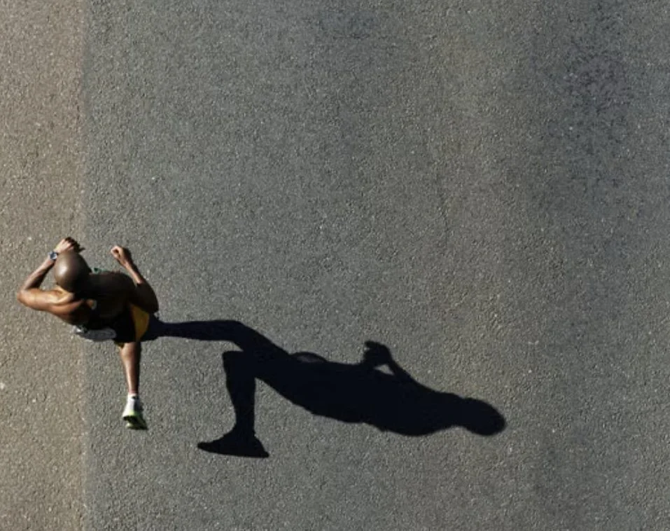 A person running on an asphalt road, casting a long shadow due to sunlight, seen from above.