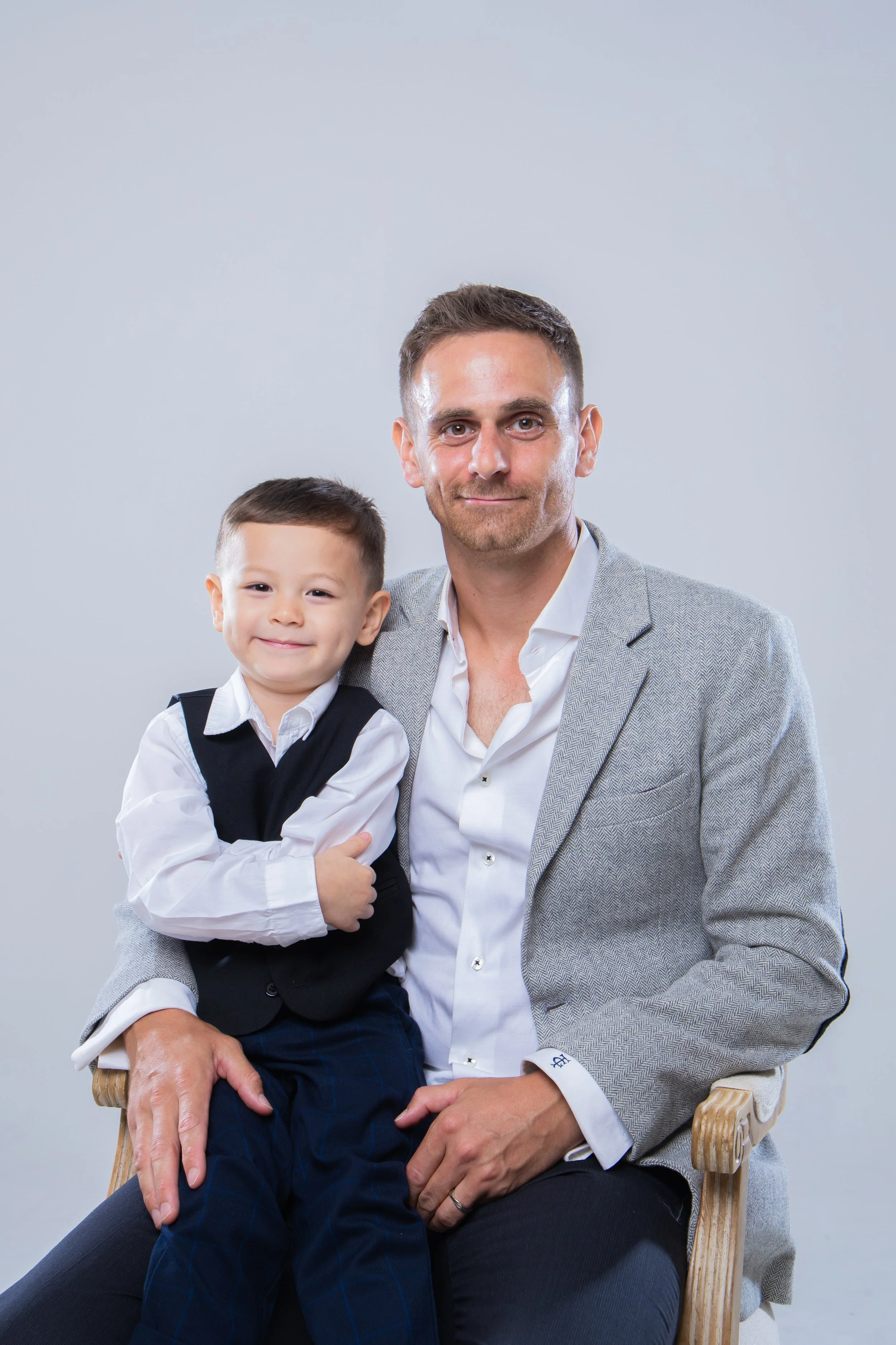 A man in a gray blazer and white shirt sitting on a wooden chair, holding a smiling young boy in a white shirt and black vest on his lap, against a light gray background.