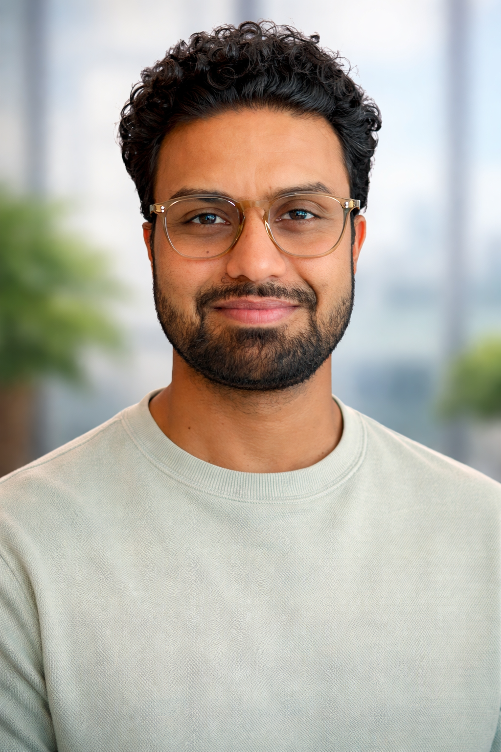 A young man with dark, curly hair, glasses, and a beard, smiling in an indoor setting with windows and blurred greenery in the background.