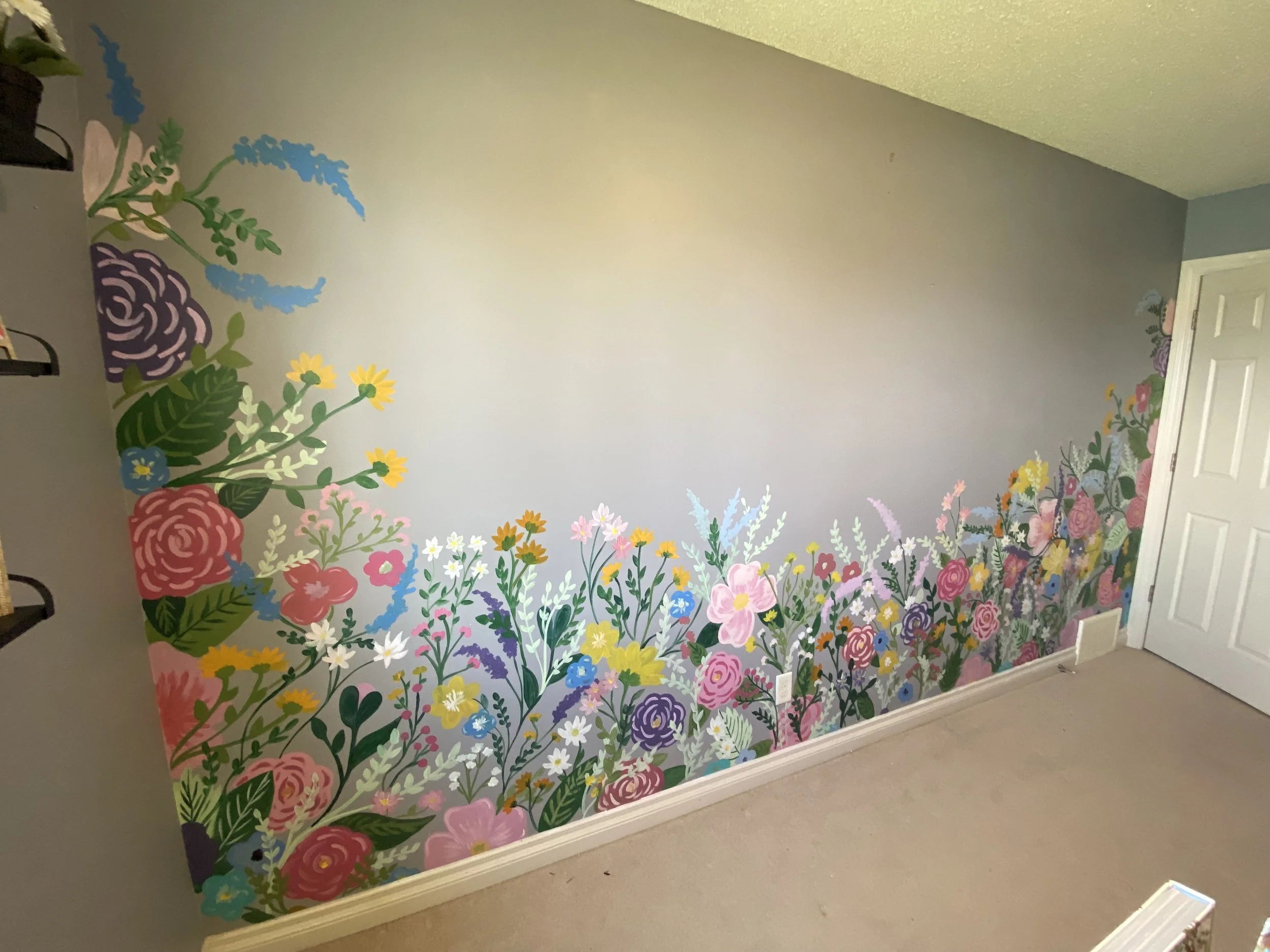 A decorated wall with a colorful floral mural featuring various flowers in pink, purple, yellow, white, blue, and green. The top of the mural includes greenery and blue accents, and it runs along the bottom of the wall up to the white door with a floral design on the upper part. The wall is painted in a neutral gray color, and the floor is beige.