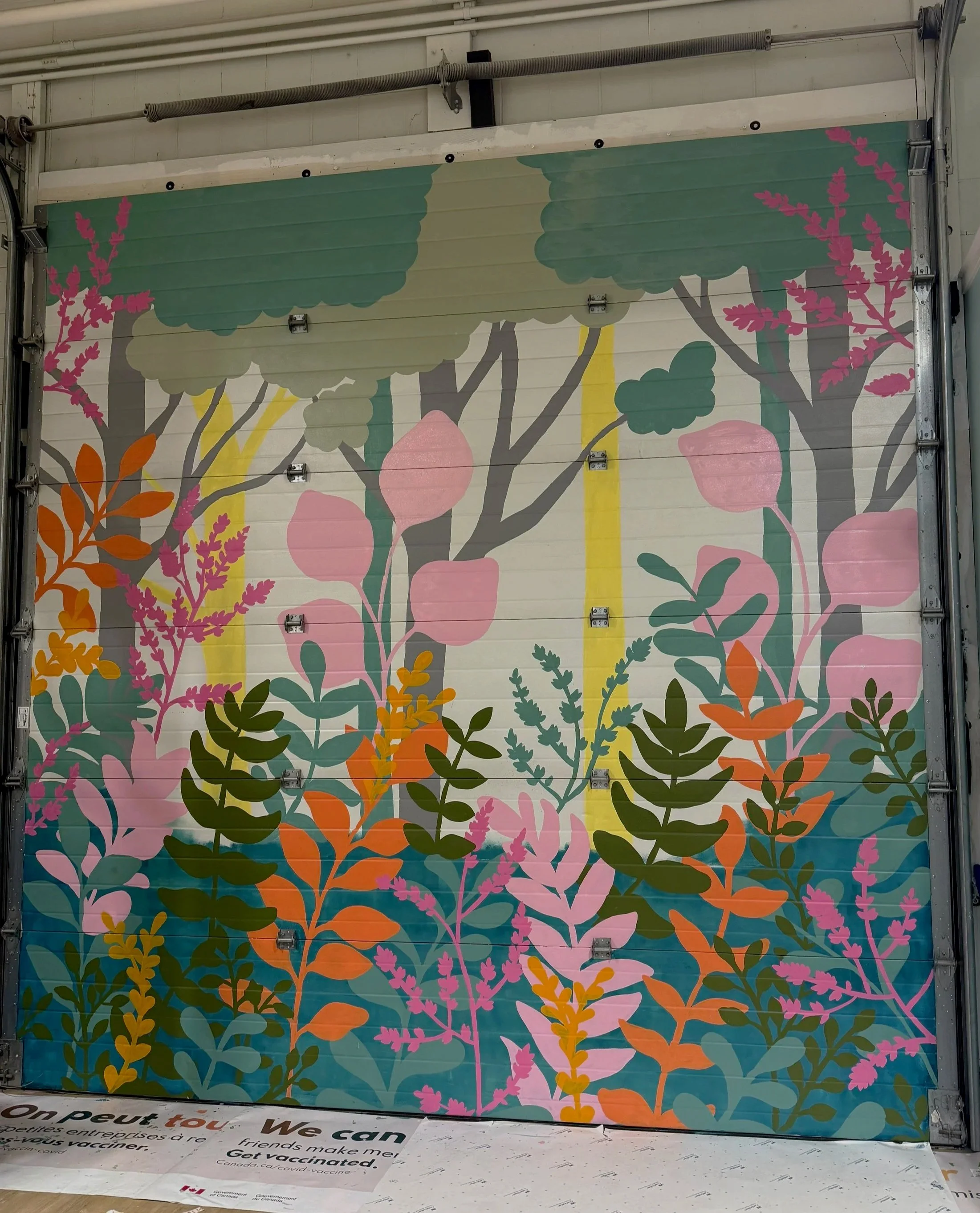 Colorful mural of stylized trees with pink, green, orange, and gray foliage on a garage door, with a partially visible sign at the bottom promoting COVID-19 vaccination in Canada.
