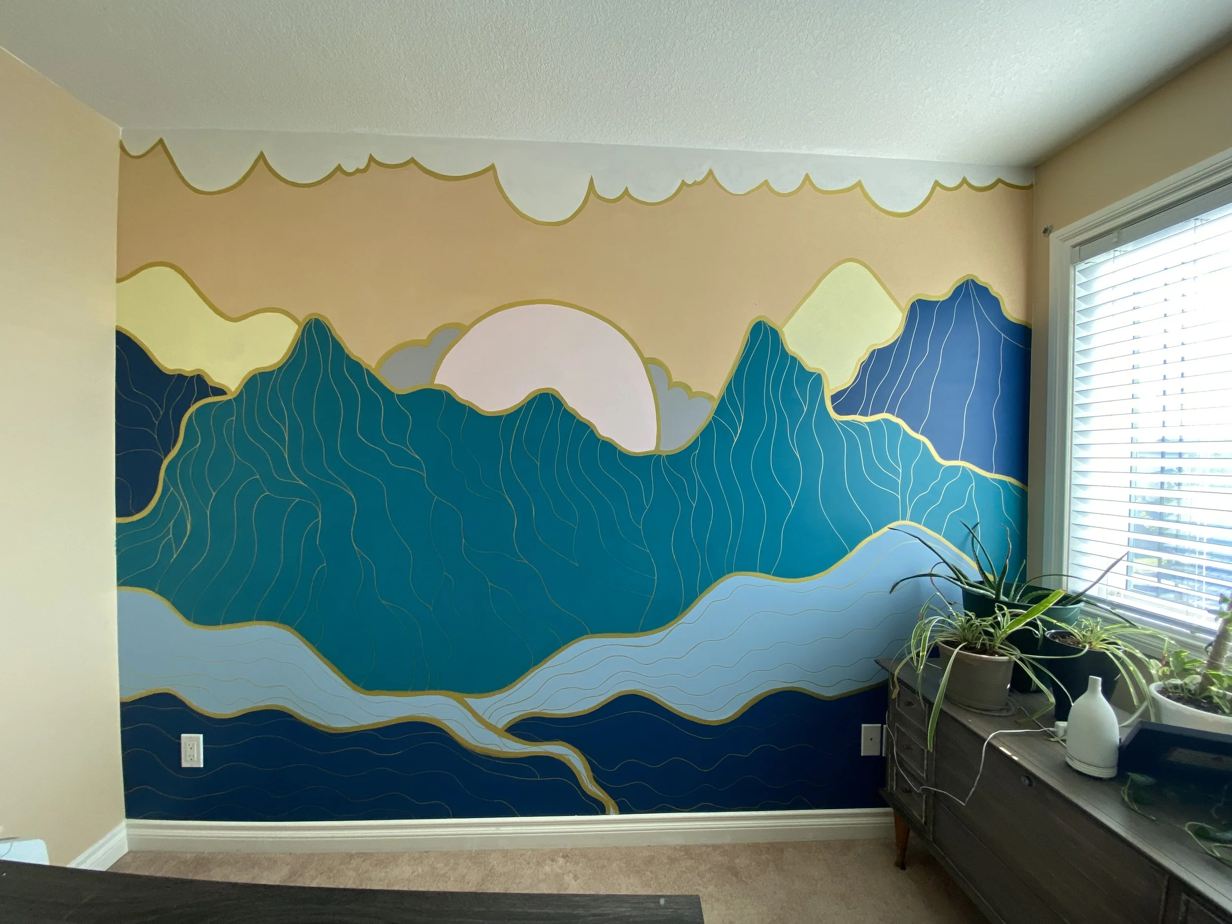 Interior wall painted with a mountain landscape mural featuring blue mountains, yellow highlights, and a pale pink sun, with a plant on a black wooden table in front of the window.