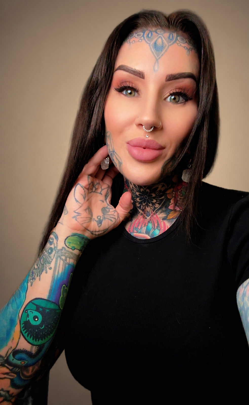 A woman with long dark hair, tattoos on her face and arms, and piercings, wearing a black shirt, looking into the camera with a neutral expression, against a plain background.