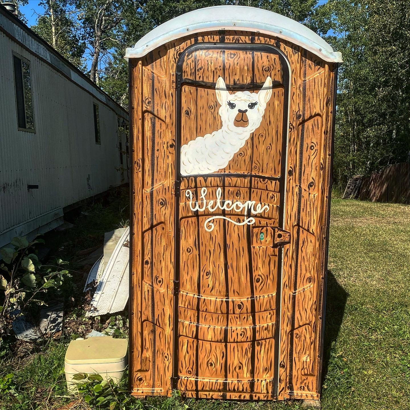 A outdoor bathroom with custom painting by Edmonton, AB artist Carly Marie Murals painting features a  wood cabin look and a llama painted on the door a 'Welcome' sign, situated in a grassy yard with trees and a mobile home in the background.