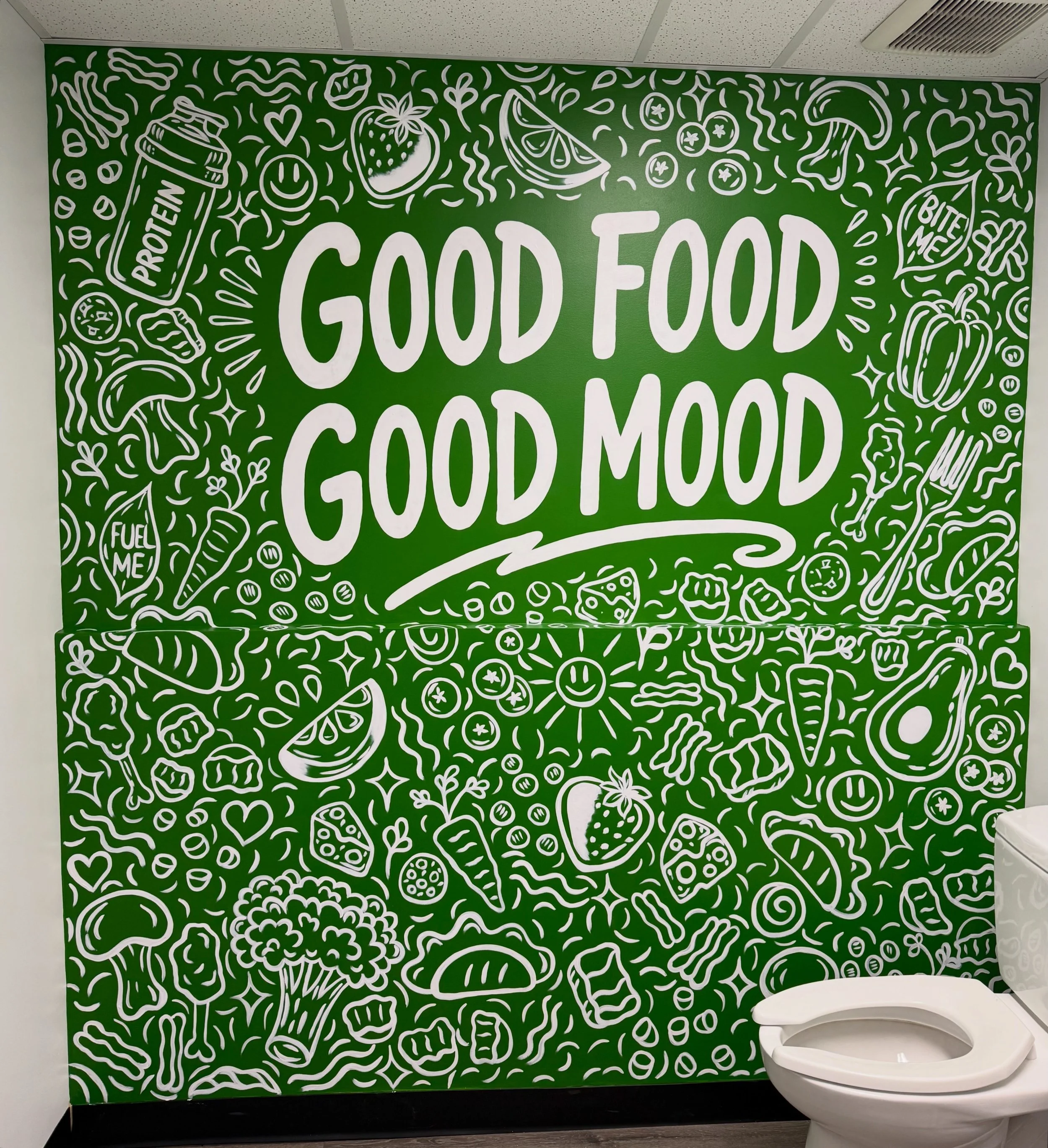 Large mural with a large green background and white hand painted food-related illustrations. The words 'GOOD FOOD, GOOD MOOD' in the center of the wall. Featured in the Just Bite Me Sherwood Park Bathroom by Carly Marie Murals