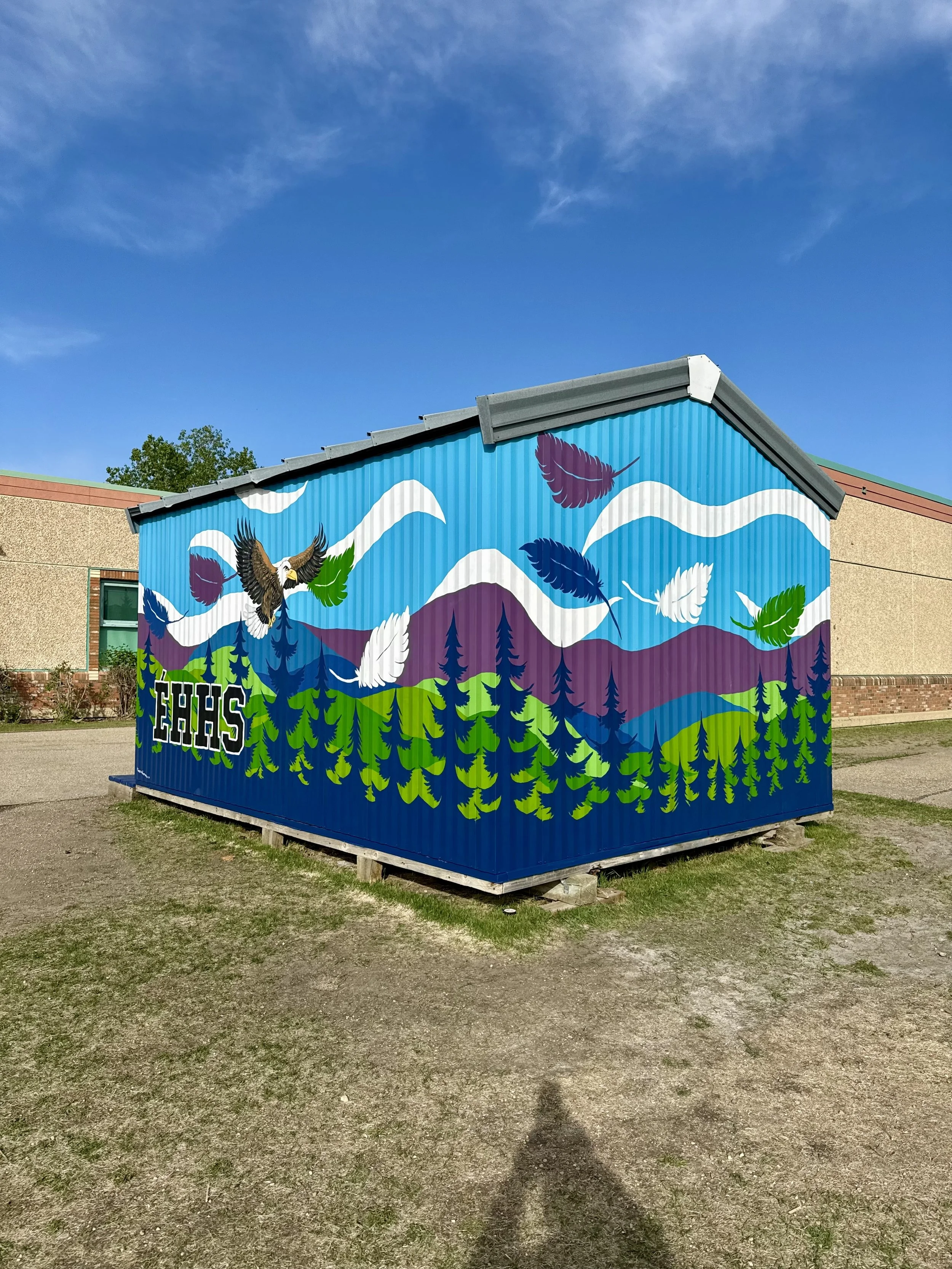 A building with a colorful mural of mountains, trees, clouds, an eagle, and feathers painted on its side. The word 'EHHS' is visible on the lower part of the mural. The sky is clear with a few clouds.