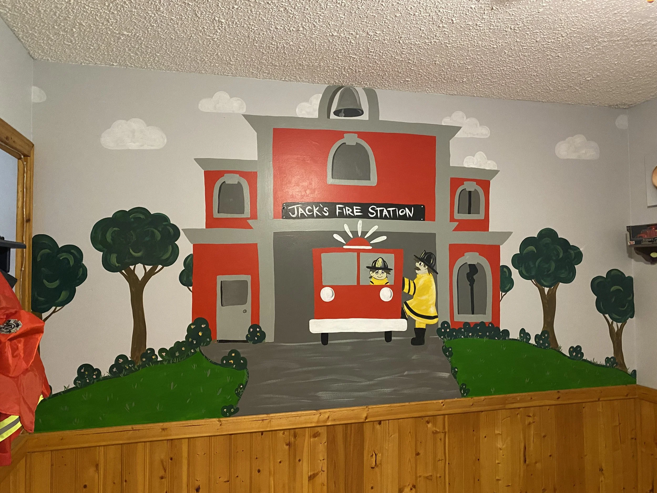 A colorful mural of a firehouse with two animated firepeople, surrounded by trees and bushes, in a children’s area of a building.