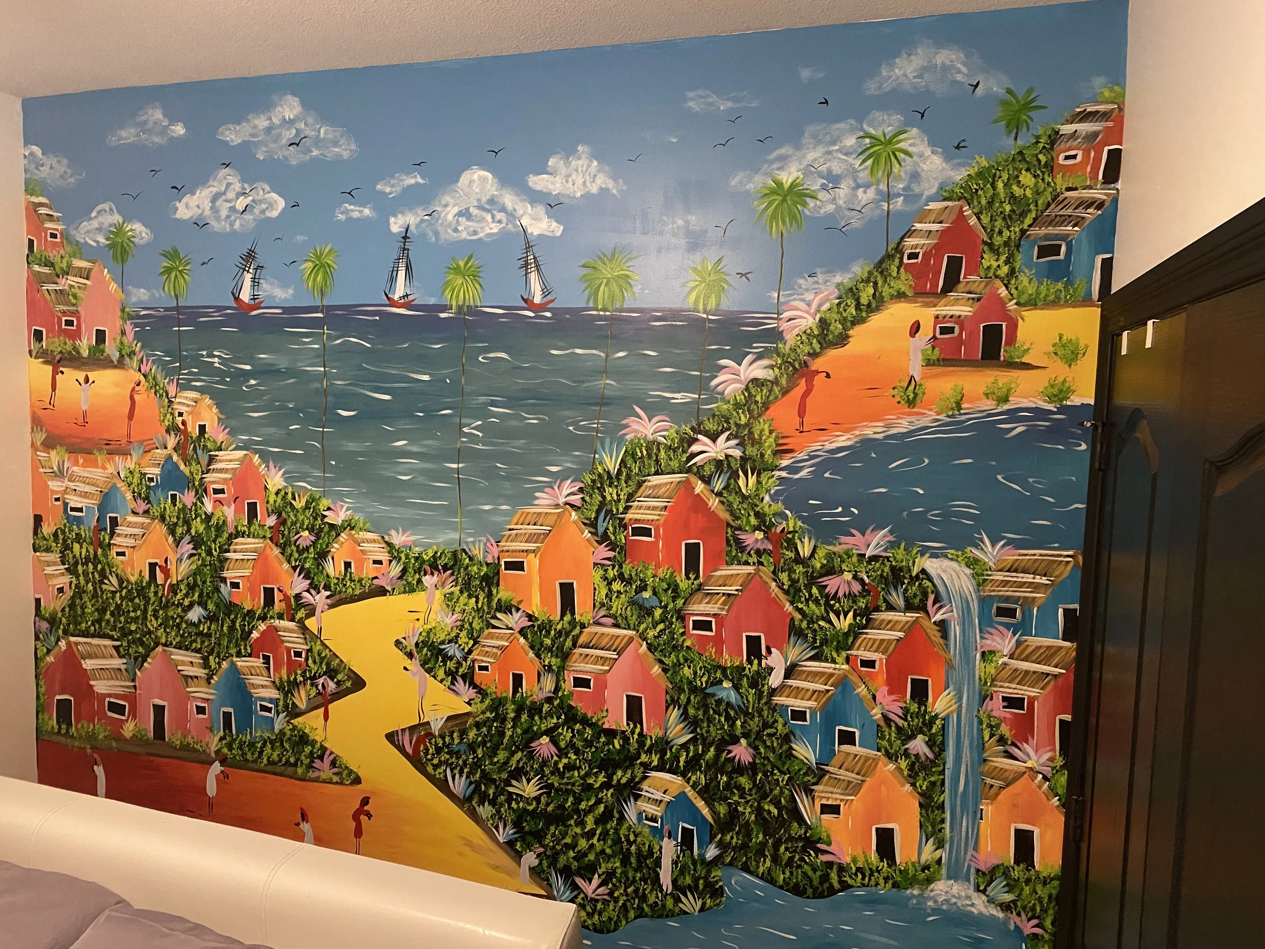 A colorful mural depicting a seaside village with pink, orange, and blue houses, lush green trees, and palm trees along a coastline with sailboats under a partly cloudy sky.
