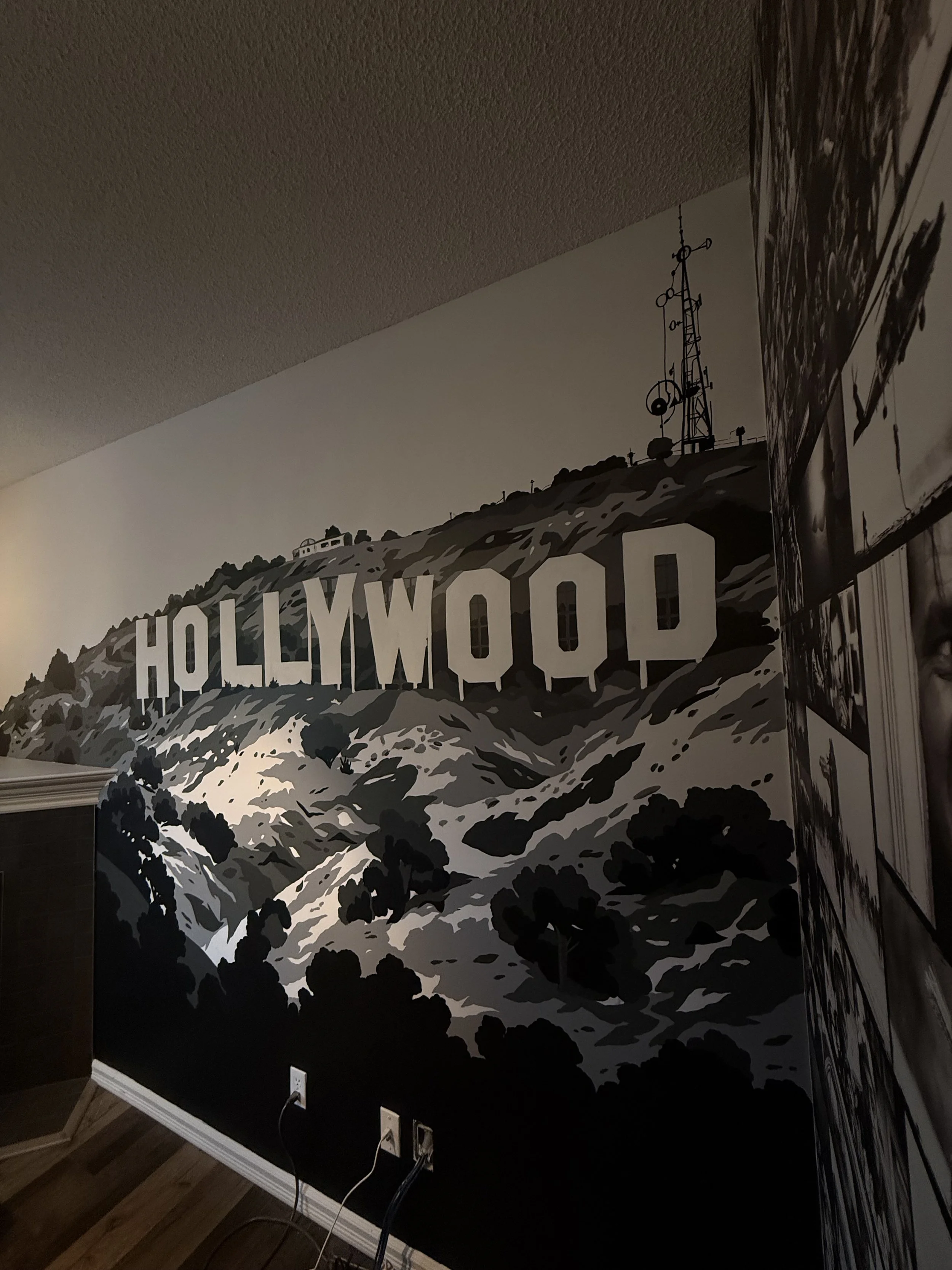 Wall mural featuring the Hollywood sign on a mountain with a communication tower, painted in grayscale tones. Hand painted in residential home featured in their theatre room by Carly Marie Murals.