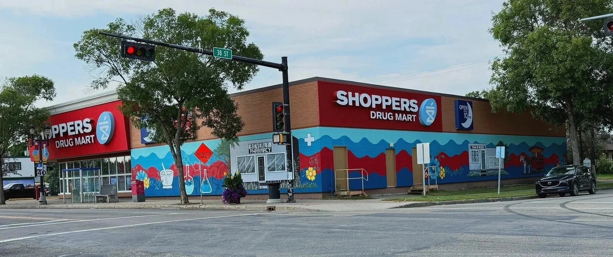 A colorful building with a mural, featuring the sign 'Shoppers Drug Mart' and a logo, situated at an intersection with traffic lights and trees on a cloudy day.