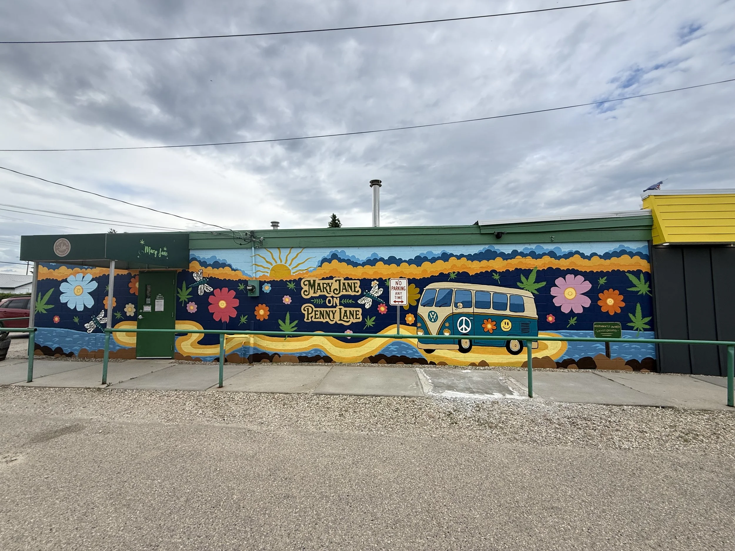 Colorful mural on building wall featuring flowers, a butterfly, a sunflower, clouds, and a Volkswagen van with peace signs, with the text "Mary Jane on Penny Lane" and various green leaves and cannabis motifs.