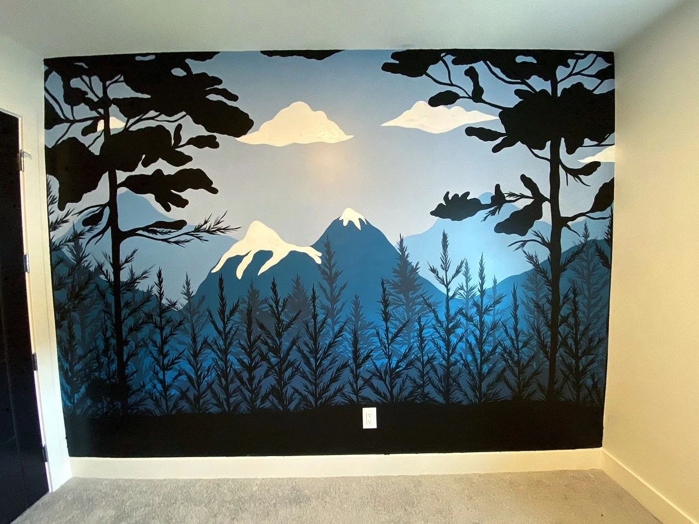 Wall mural depicting mountain landscape with snow-capped peaks, pine trees, clouds, and a blue sky, framed by silhouettes of trees in black.