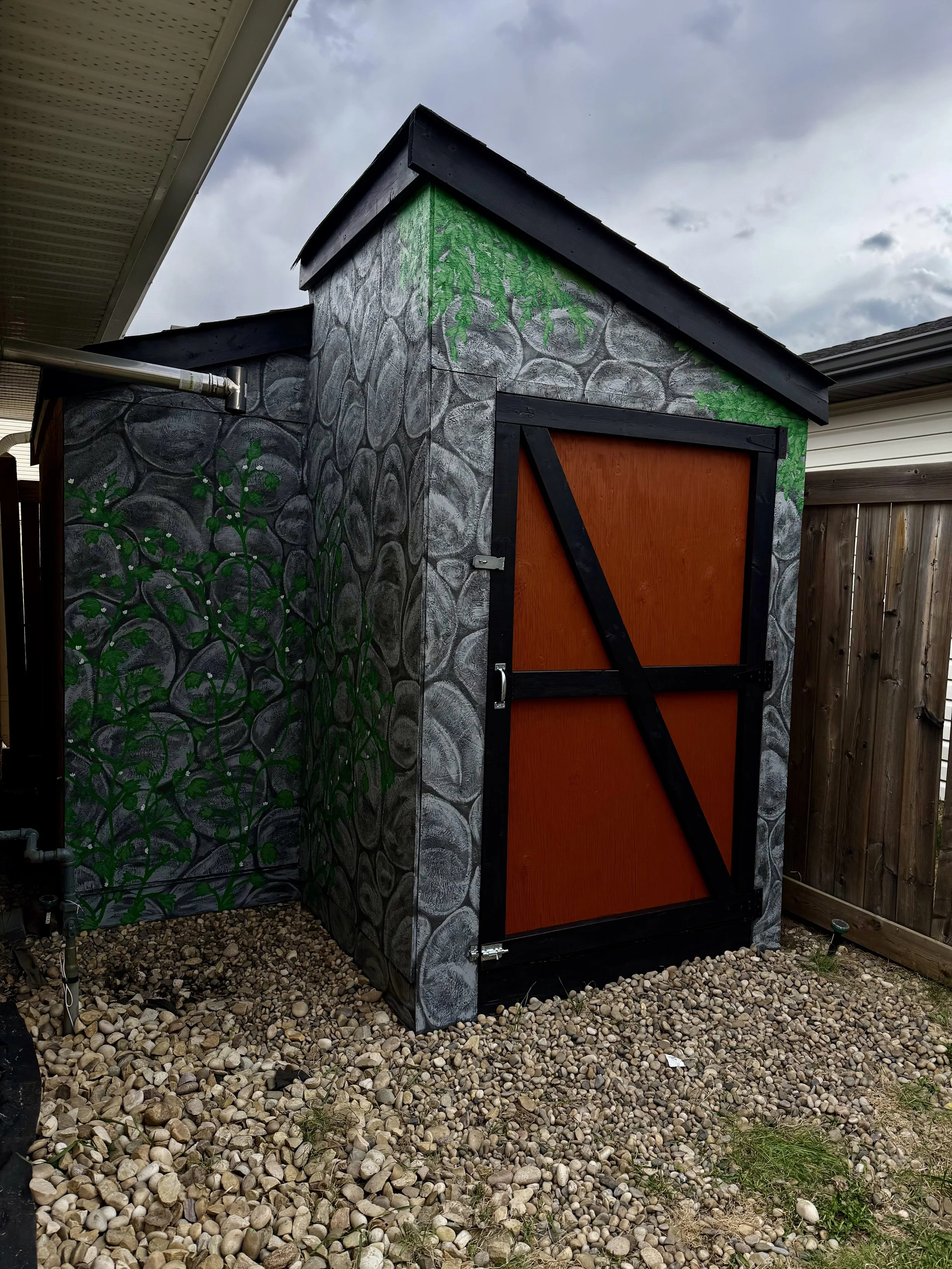 A small outdoor shed with a stone-patterned exterior, painted green with black trim and a large, orange door. The shed is situated on a gravel yard, and part of a neighboring fence is visible on the right side. The sky above is cloudy.