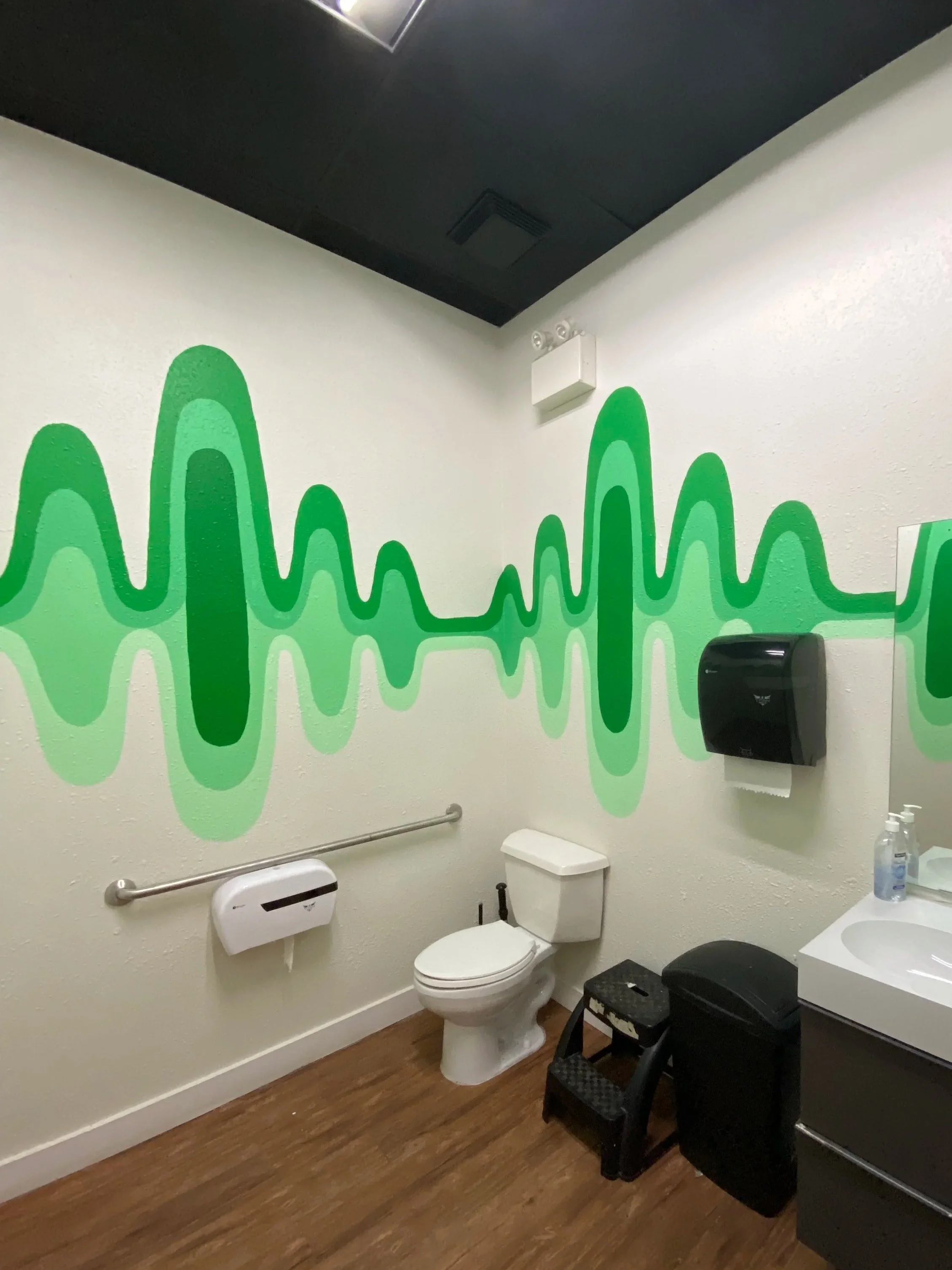 A bathroom with a white toilet, a black trash can, wall art with green wave patterns, a mirror, soap dispenser, and wooden floor.