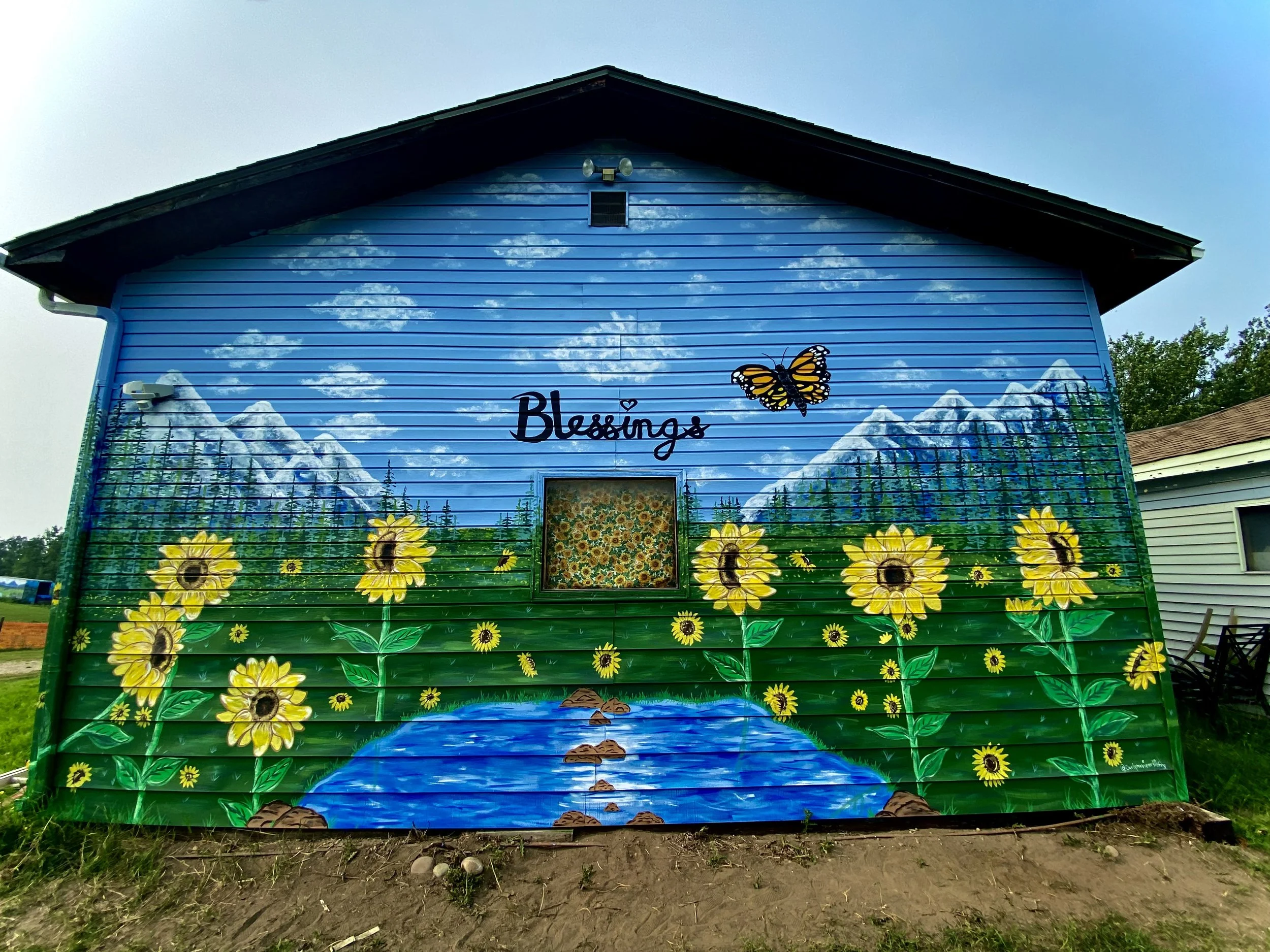 A colorful mural of a mountain landscape with a flowing river, sunflowers, and a butterfly on a blue house exterior wall with the word 'Blessings' written.