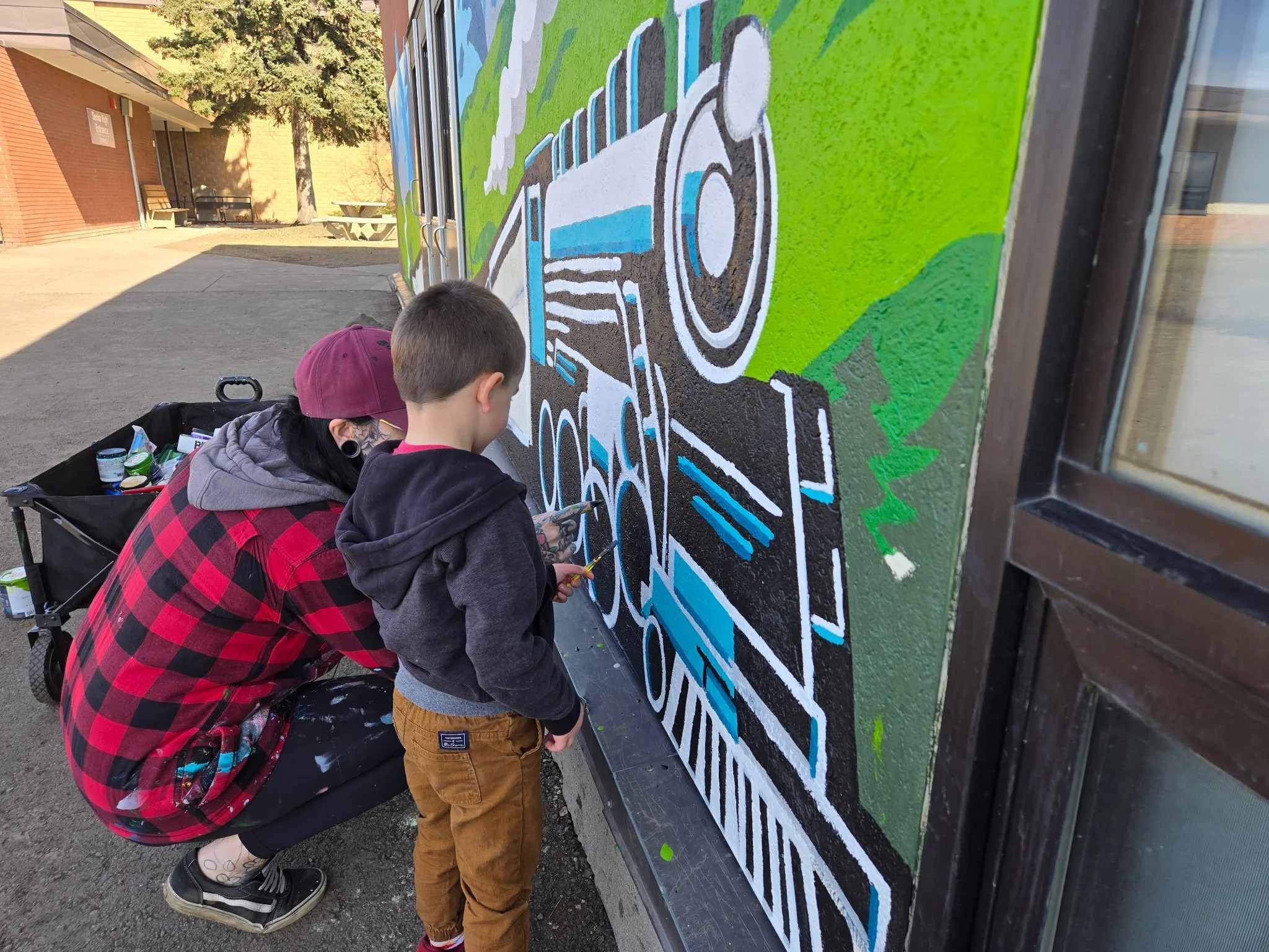 Two boys, one with a maroon cap and a tattoo on his arm, and the other with brown pants, are painting a large mural on a wall outdoors. The mural features a train with a bright green background and white, black, and blue details. It appears to be part of a community or school project.