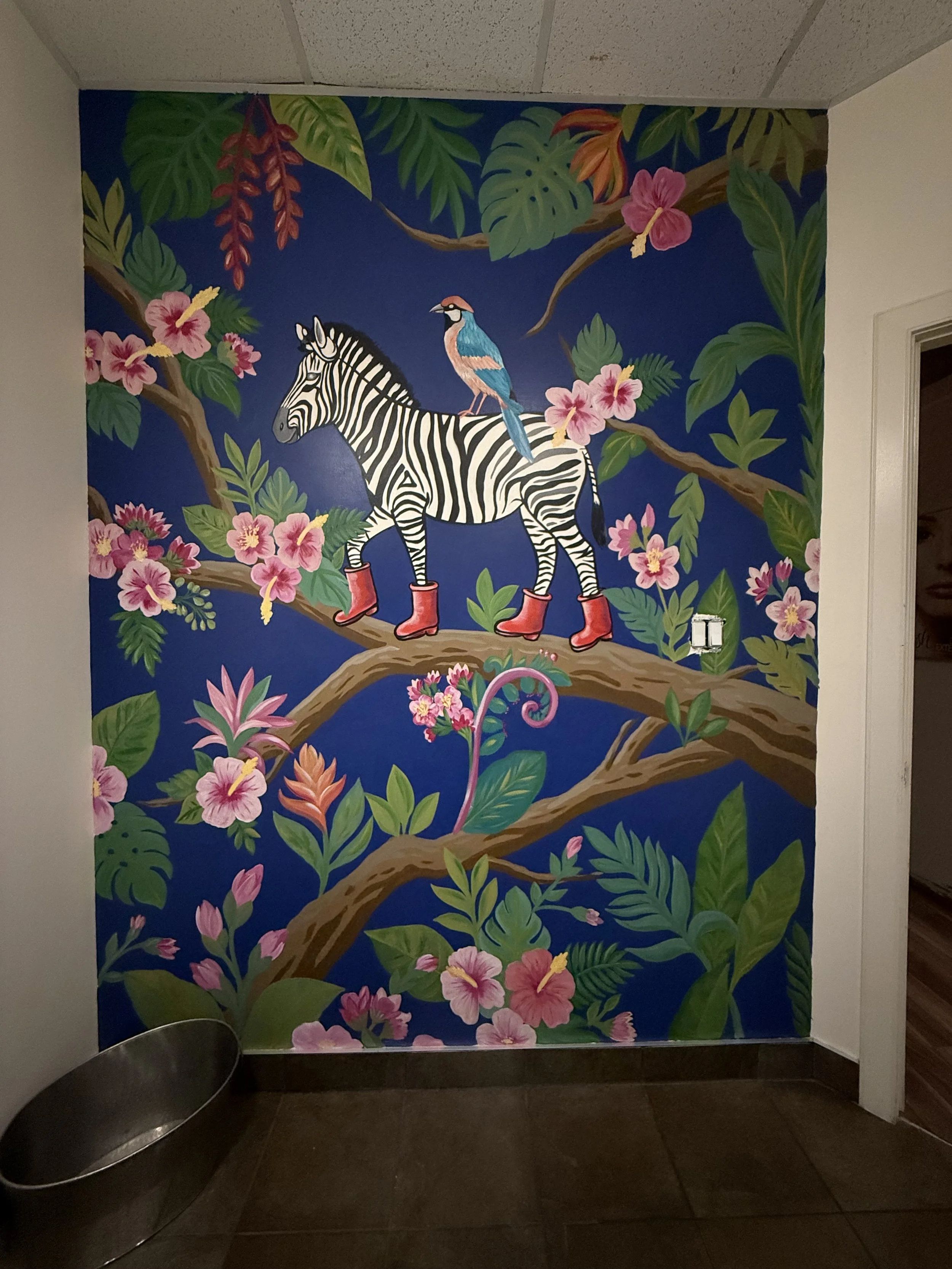 Colorful mural feature wall in Fubutan bathroom depicting a zebra with a bird on its back, surrounded by lush green leaves and pink flowers on a blue background. Hand painted by Carly Marie Murals