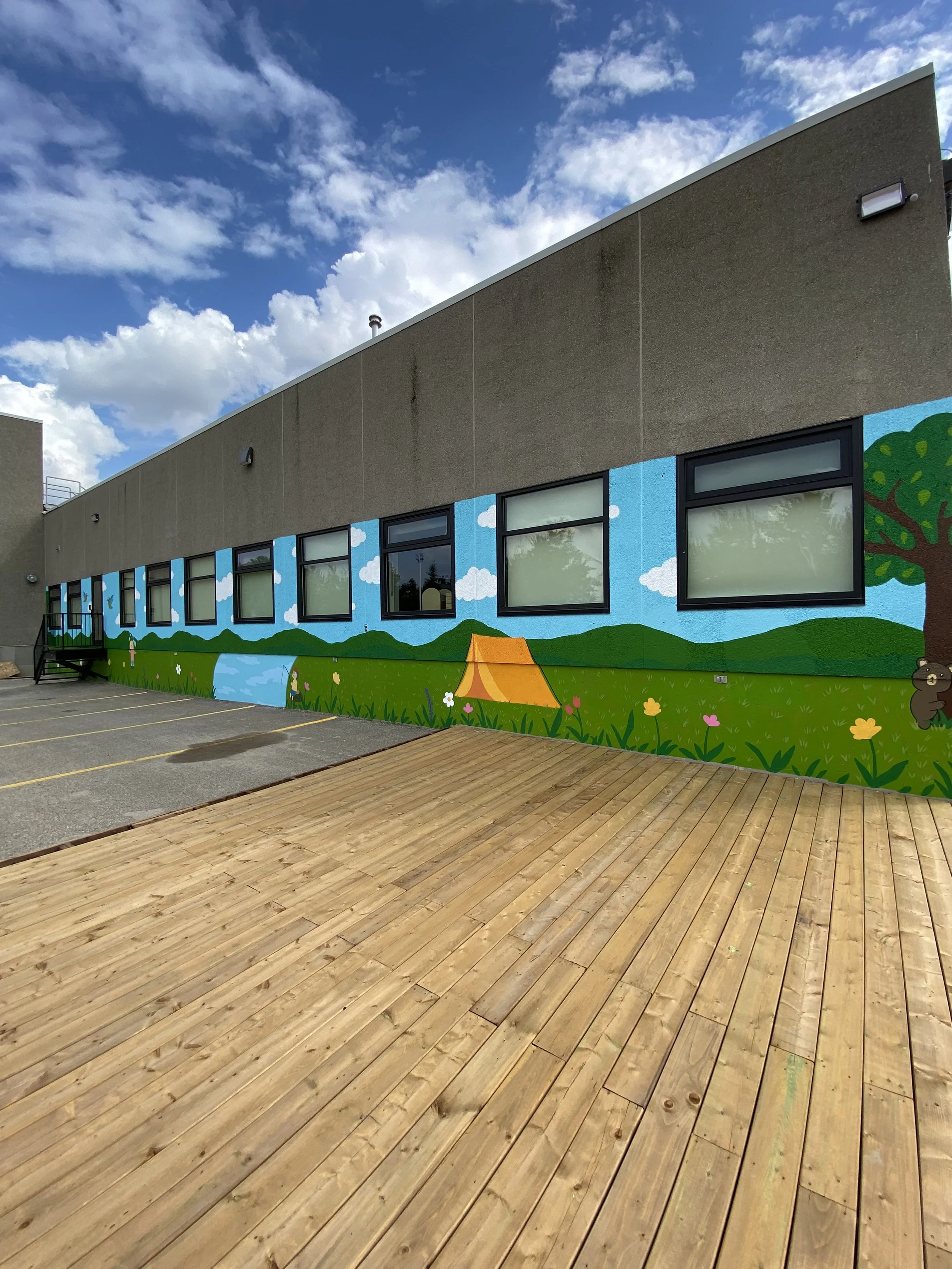 A building with a mural of clouds, trees, and grass painted on the exterior wall. There are windows, some with reflections, and parking space lines on the asphalt in front. The sky above is blue with white clouds.