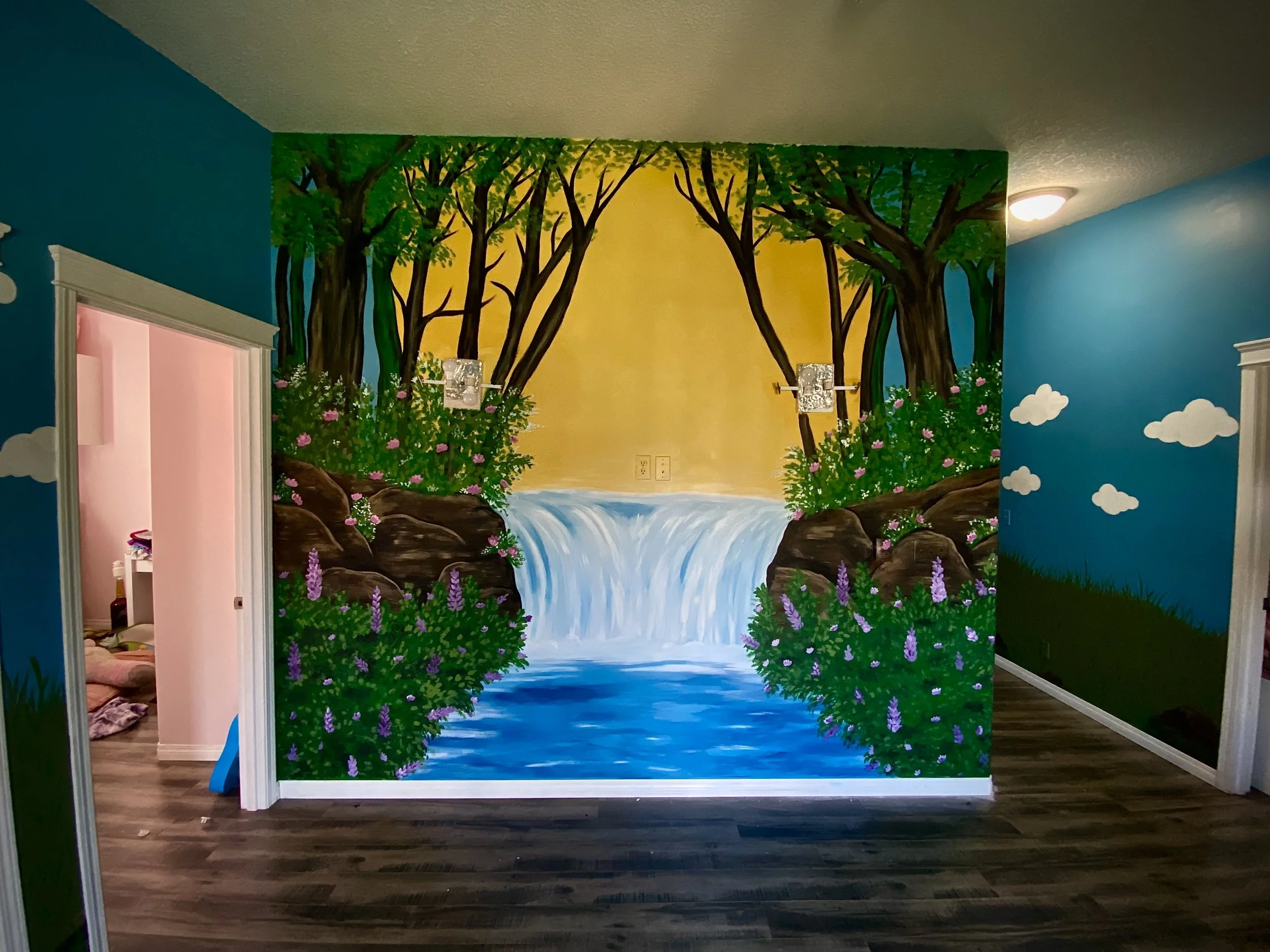 Room with a large mural painted on the wall depicting a nature scene with trees, a waterfall, and water, surrounded by painted plants and clouds.