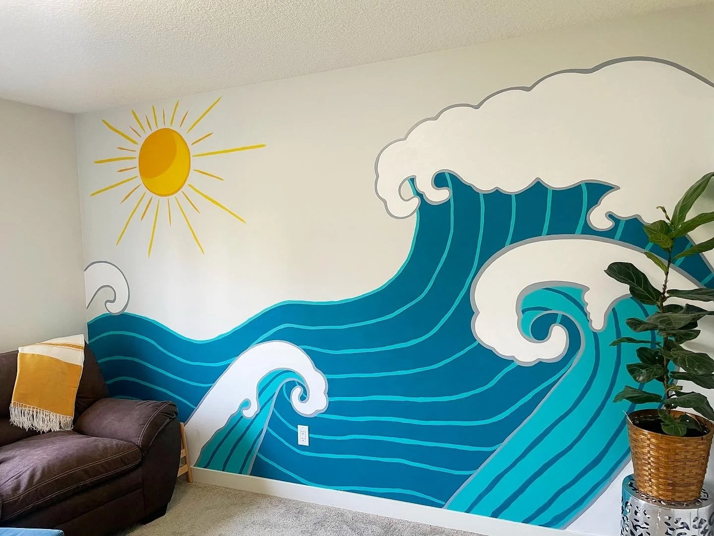A wall painted with a mural of a large blue ocean wave, a yellow sun with rays, and a large white cloud, in a living room with a brown couch, a yellow and white blanket, and a potted plant.