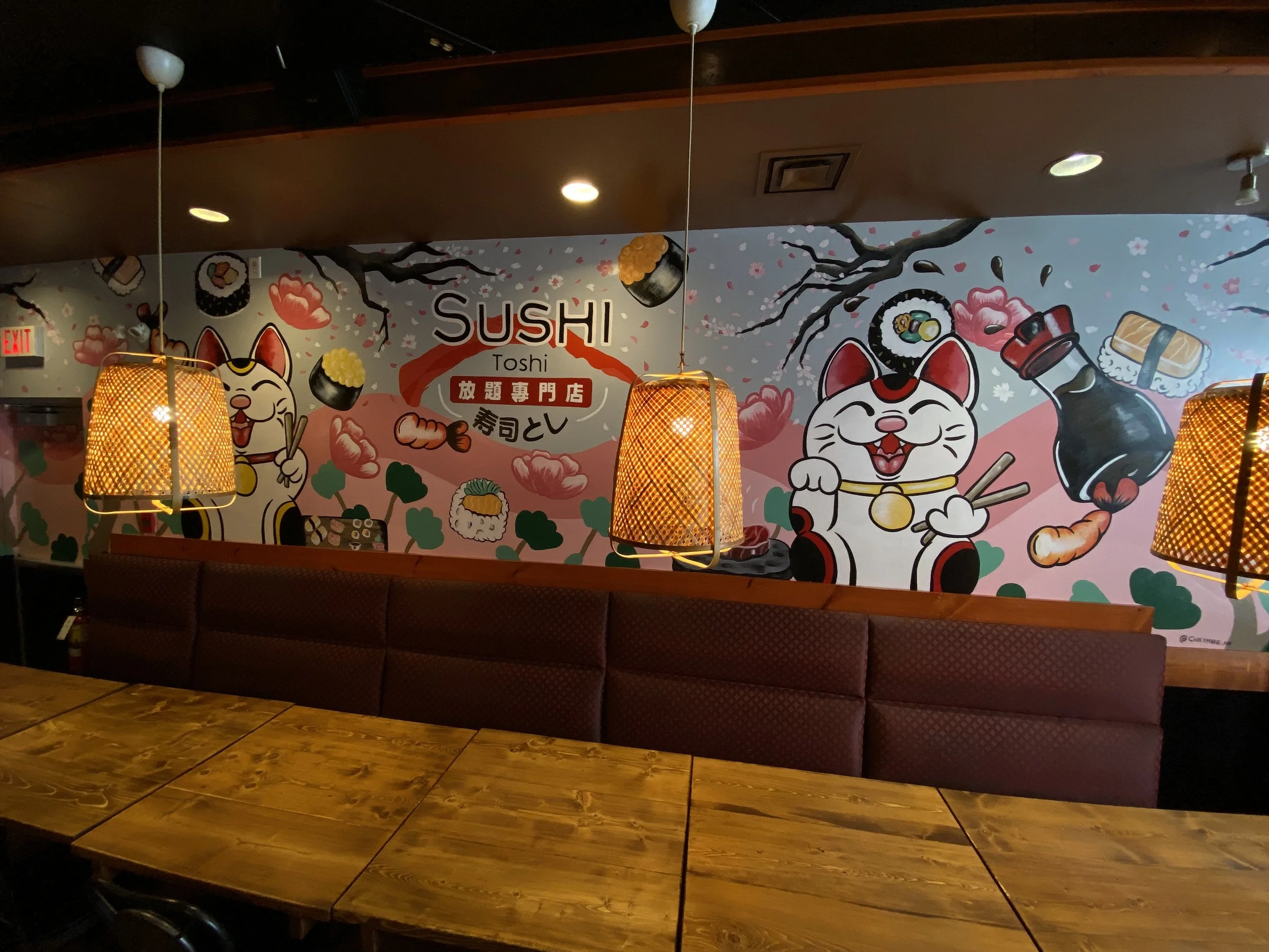 Decorative wall mural in a restaurant featuring cartoon cats with sushi, chopsticks, and sake bottles, with hanging orange lamps and wooden tables in the foreground.