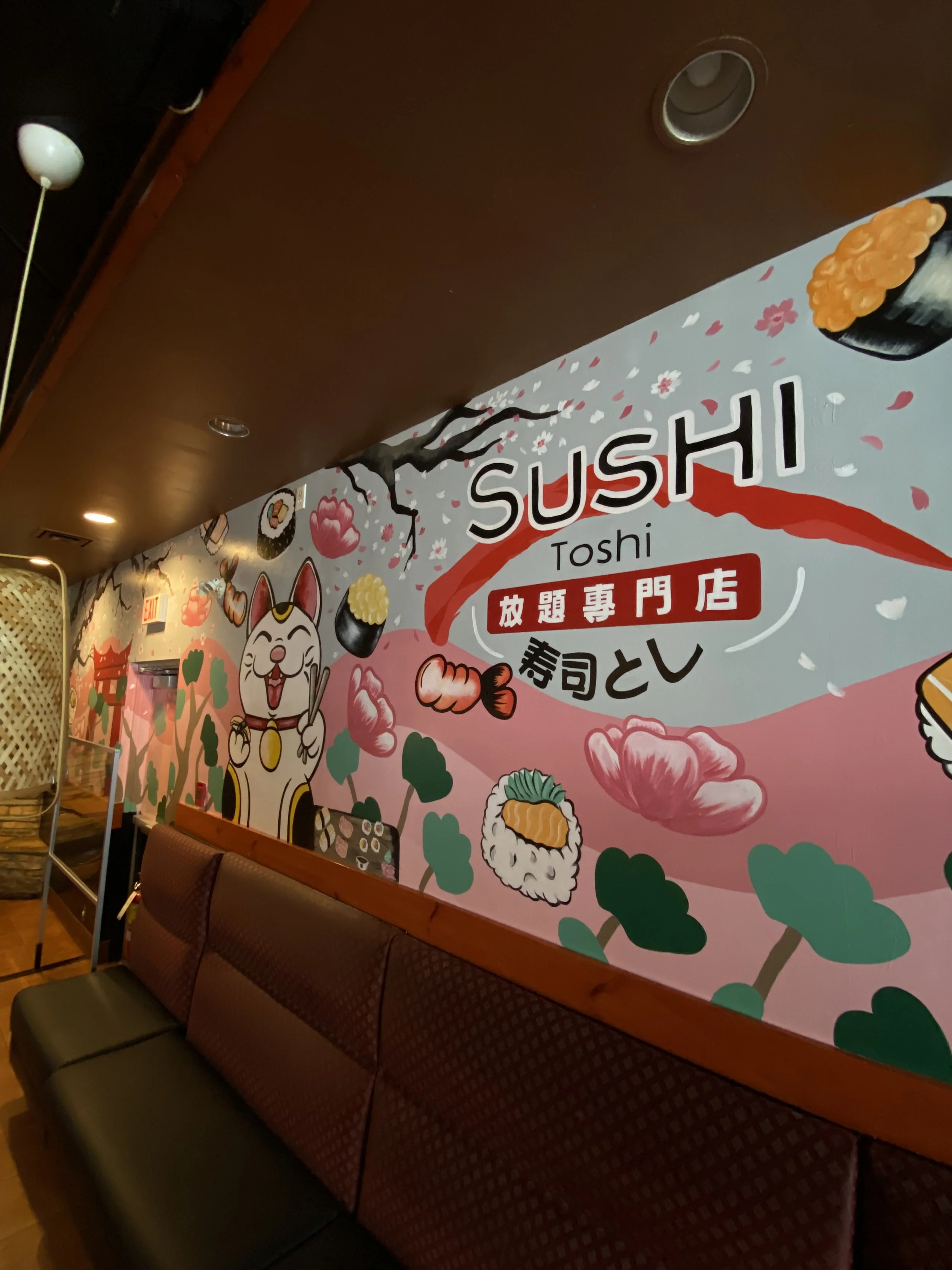 Interior of a sushi restaurant with a mural on the wall featuring sushi, cherry blossoms, a lucky cat, and Japanese-themed illustrations. A row of cushioned seats lines the wall.