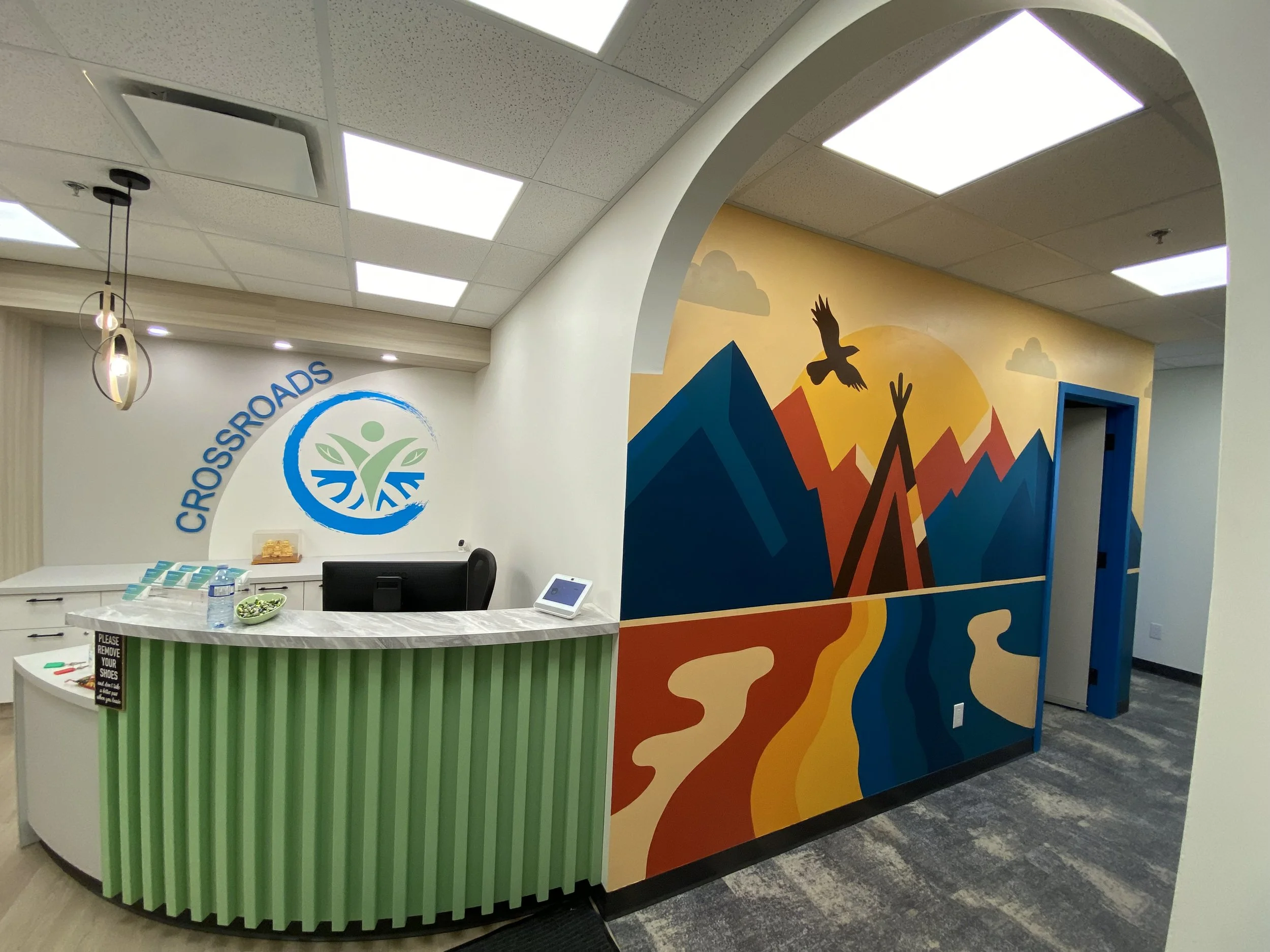 Reception desk in a health or wellness facility with a green striped front, a computer screen, and a chalkboard sign. Behind the desk is a wall with a colorful mountain, sun, clouds, and flying birds mural, and a blue door to the right.