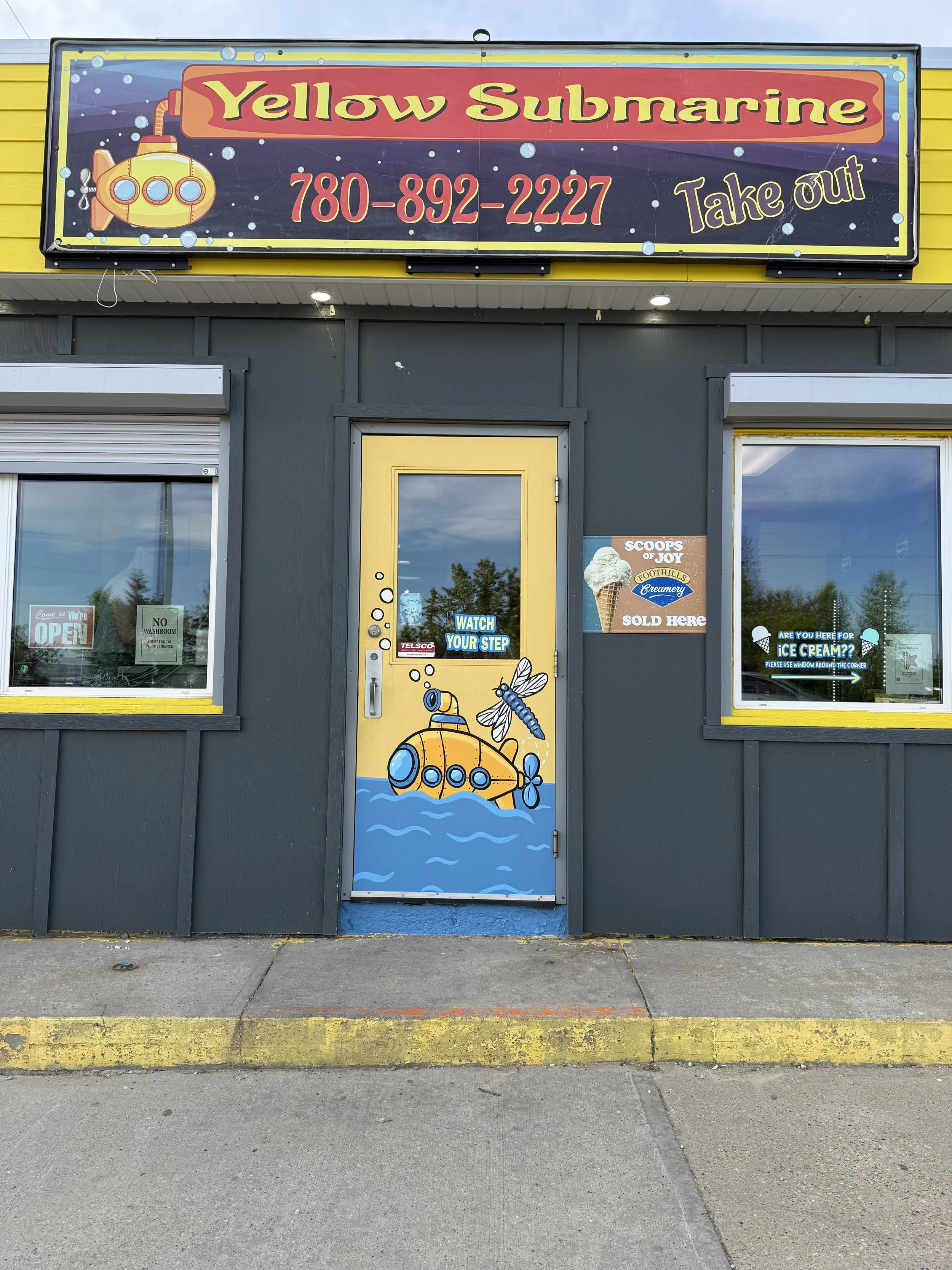 Sign for Yellow Submarine restaurant or store with a yellow and red sign displaying the phone number 780-892-2227 and the words 'Take out.' The door and windows are decorated with underwater theme images, including a yellow submarine, an ice cream cone, and a bee. The building exterior is painted dark gray with yellow accents.