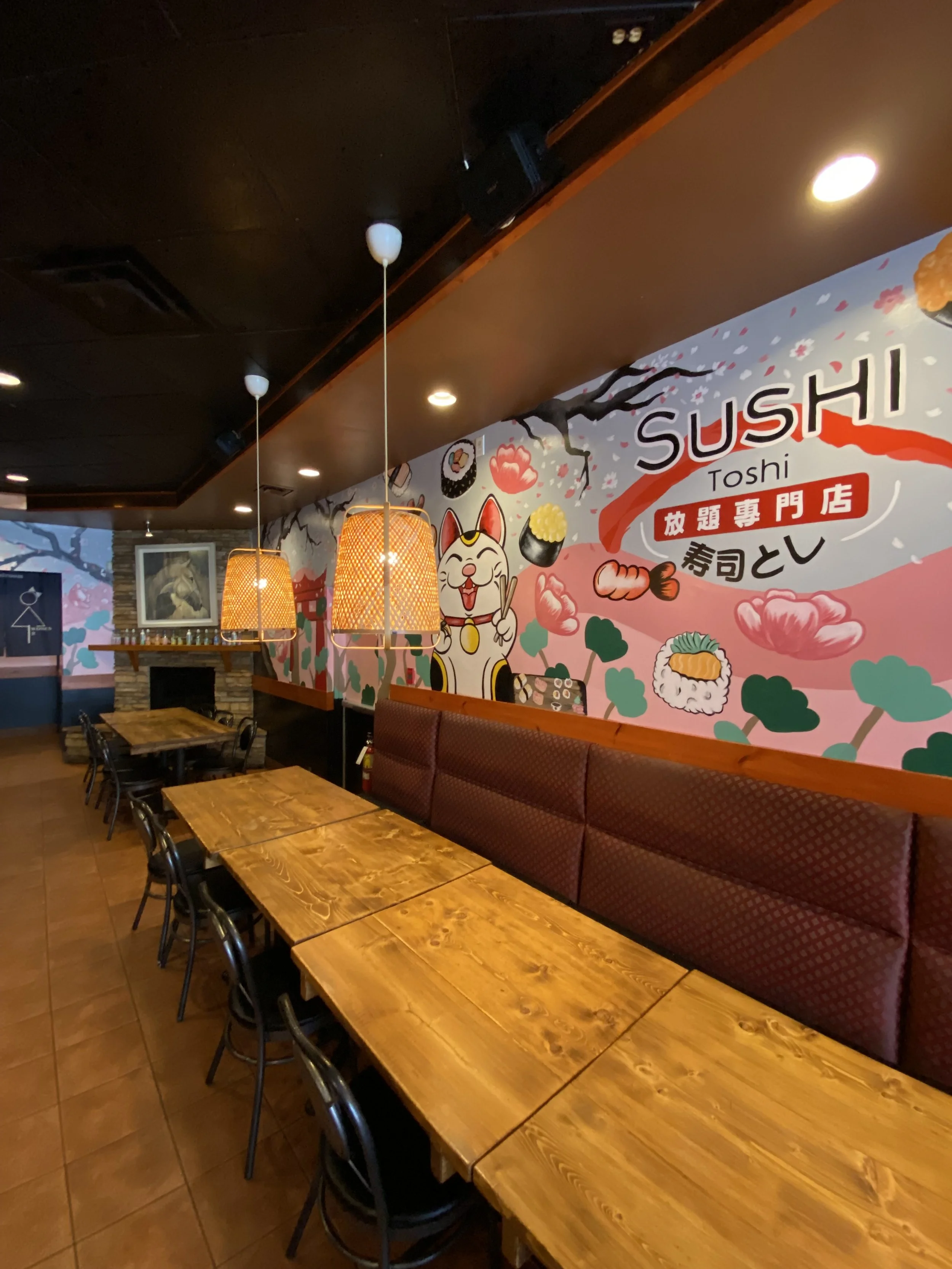 Interior of a Japanese sushi restaurant with wooden tables, black chairs, a maroon upholstered bench, pendant lights, and a colorful mural wall featuring colorful illustrative beckoning lucky cat, sushi, and Sushi Toshi Logo by Edmonton artist Carly.