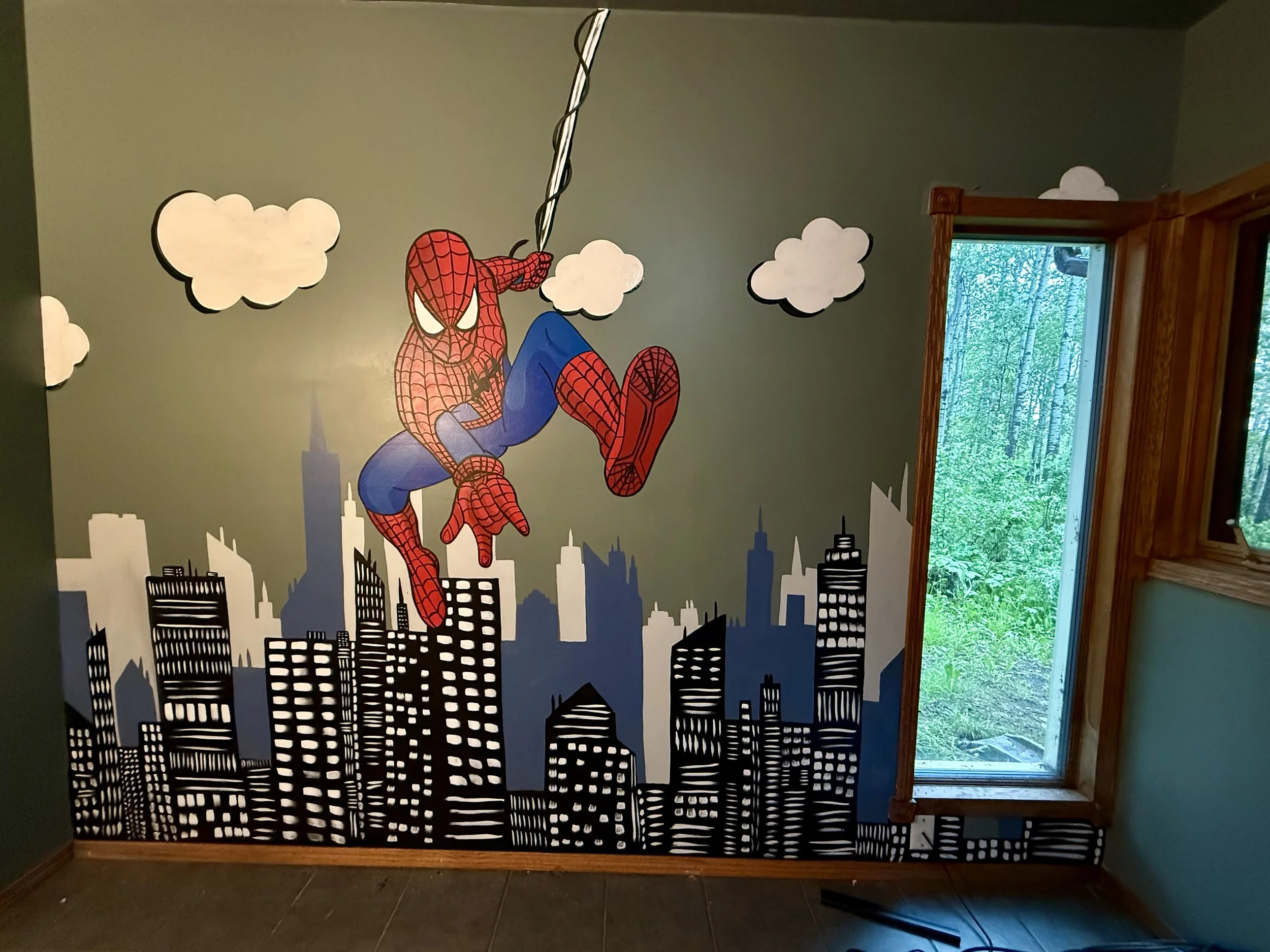 A wall mural featuring Spider-Man swinging over a city skyline with clouds in the background in a children's kids bedroom mural by Edmonton, AB artist Carly Marie Murals