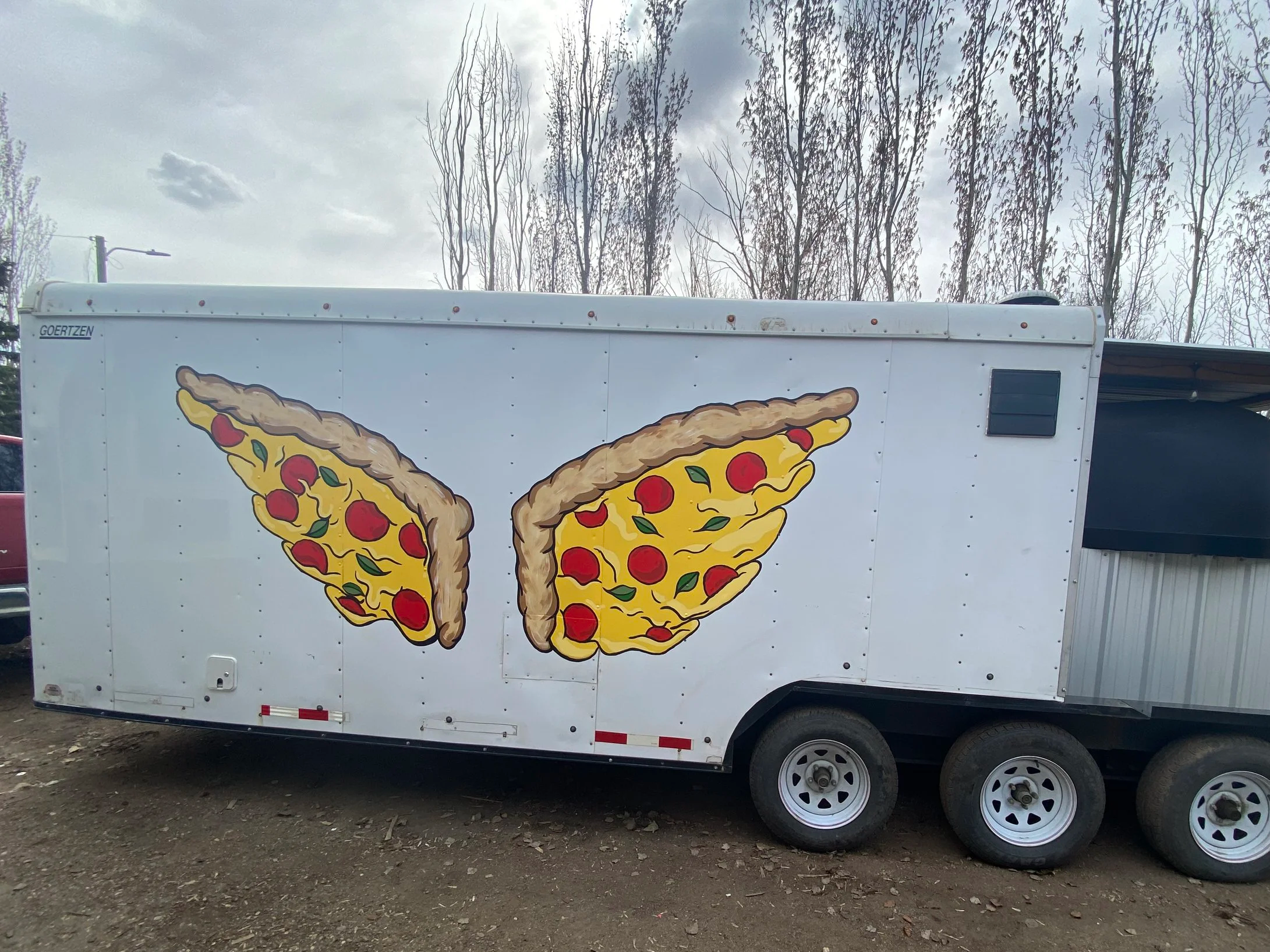 Food truck decorated with large pizza slice graphics on the side, parked outdoors with leafless trees and cloudy sky in the background.