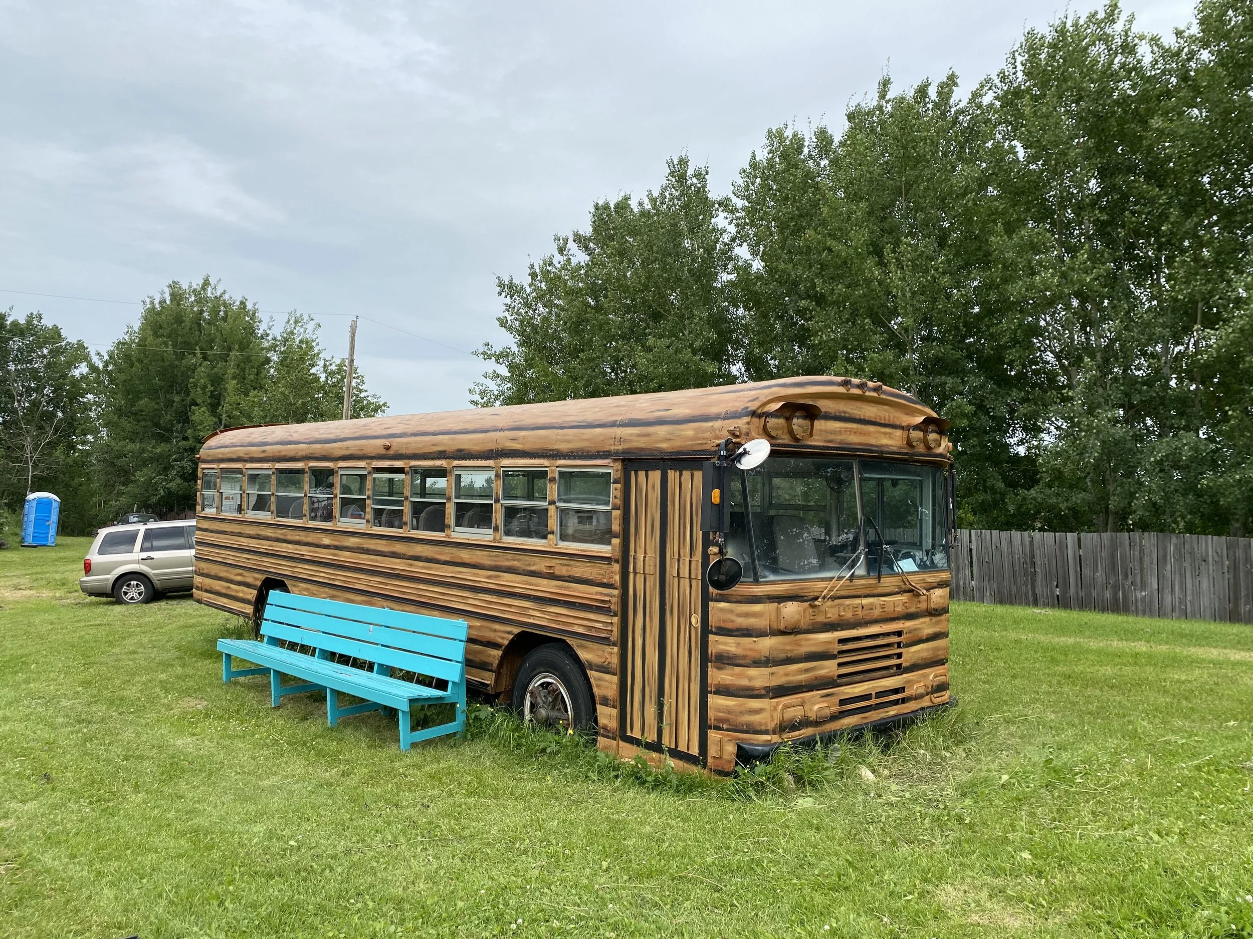 School bus parked in open field painted to look like a cabin with a teal bench and trees behind it