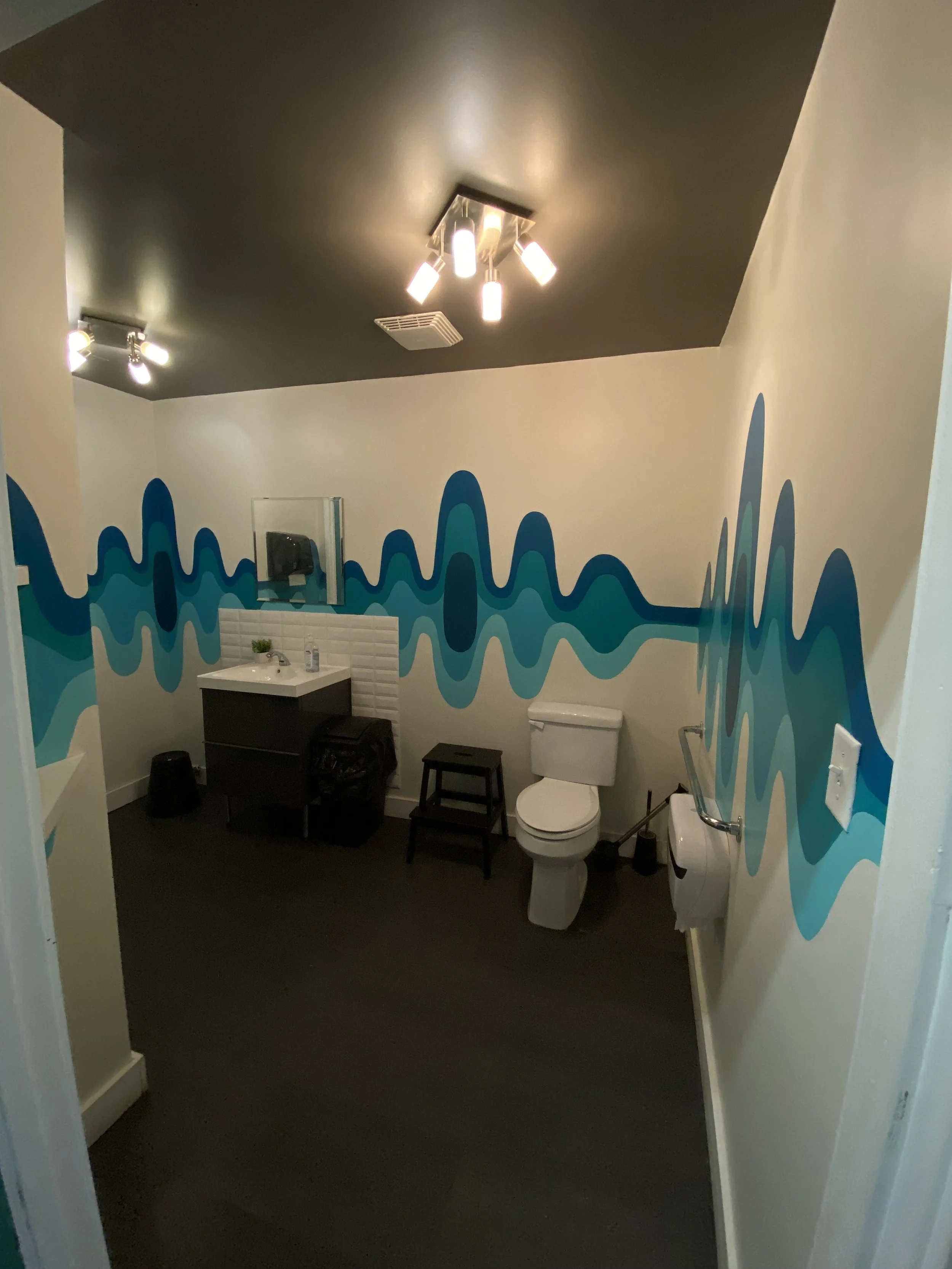A restroom with a dark ceiling, colorful wave-patterned wall, and white fixtures including a toilet, sink, and a mirror. There are multiple light fixtures on the ceiling.