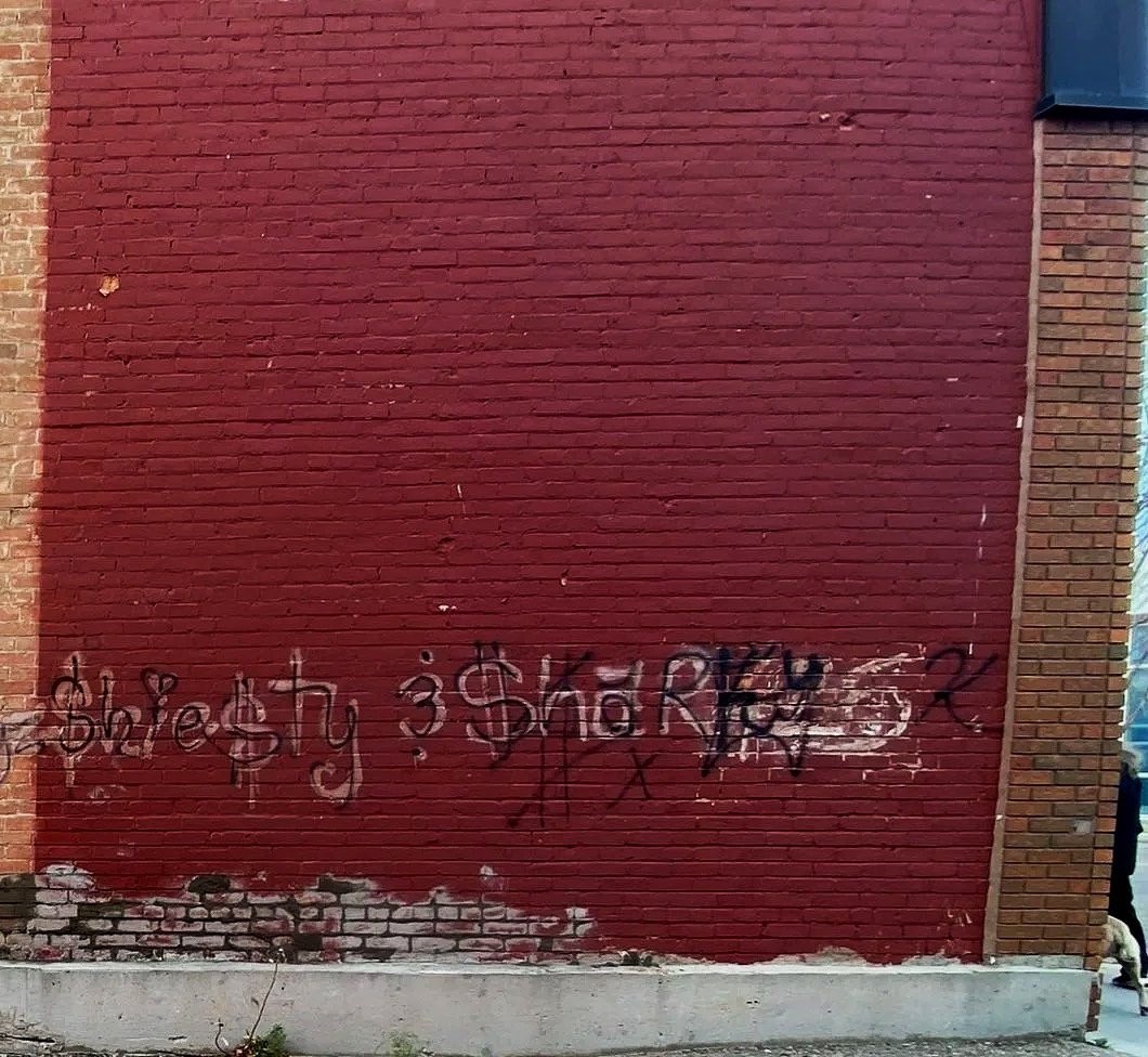 Graffiti on a red brick wall with partially faded and spray-painted tags and symbols before mural transformation by Carly Marie Murals at the Wetaskiwin Heritage Museum