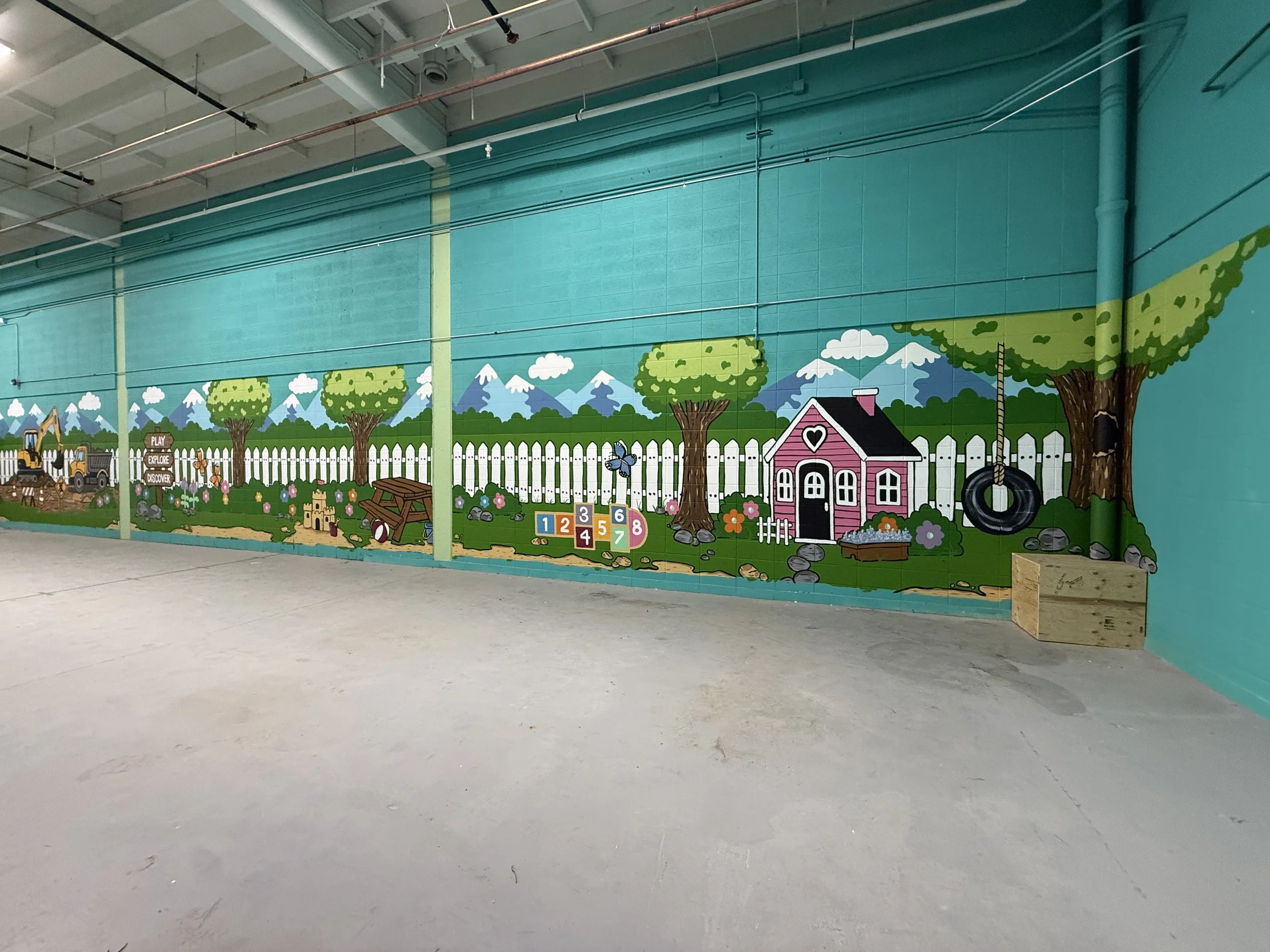 A colorful mural depicting a whimsical outdoor scene with mountains, trees, a pink house with a heart-shaped window, a tire swing, and playful animals, illustrating a child's imaginative landscape by Carly Marie Murals at indoor sandbox Edmonton, AB