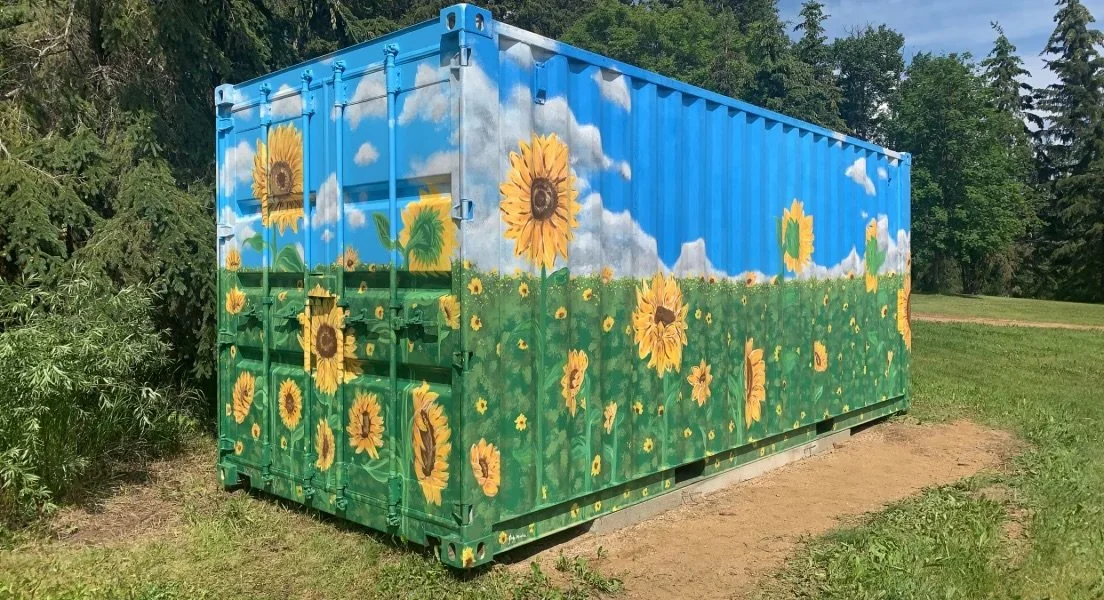 Shipping container custom painted for Country Blossoms Greenhouse in Tawatinaw, AB with a sunflower field scene, showing large sunflowers under a blue sky with white clouds. Custom painted shipping container by Edmonton, AB artist Carly Marie Murals