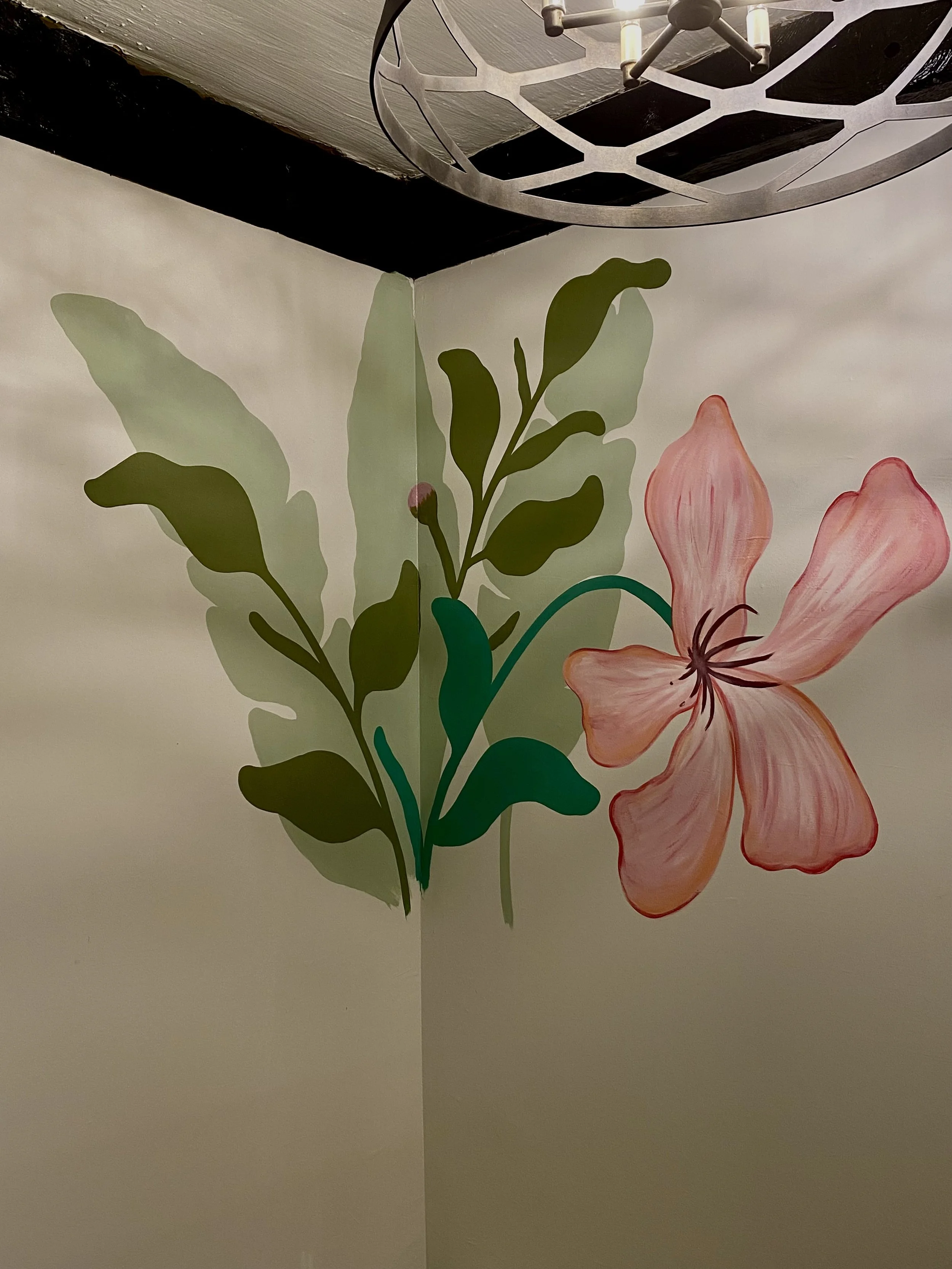A corner of a room with a wall mural of green leaves and a pink lily flower. A geometric metal chandelier with candles hangs from the ceiling.