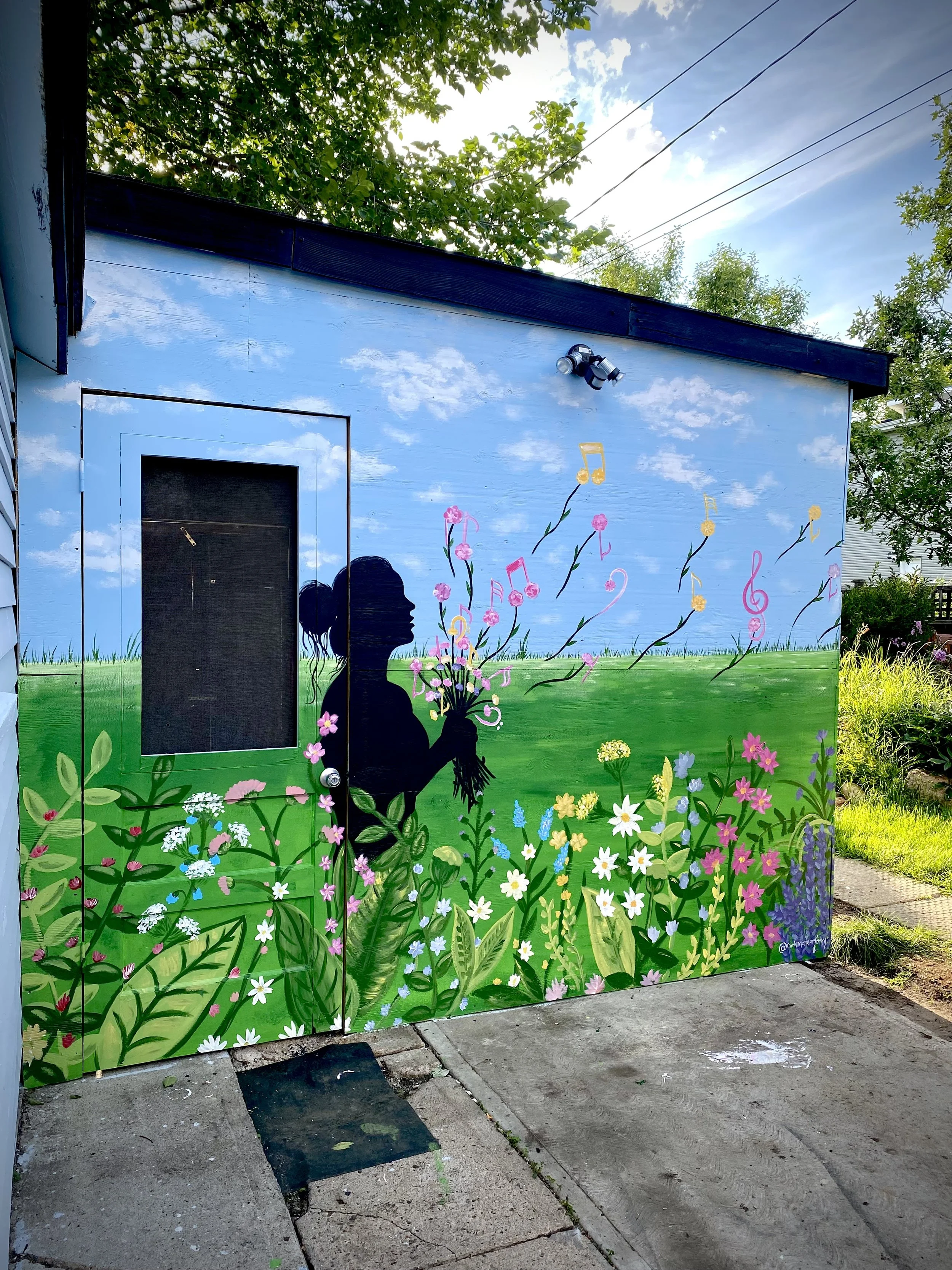 Street art mural painted on a small building depicting a black silhouette of a woman holding a bouquet of pink flowers, with a sky background, green grass, and various colorful flowers at her feet.