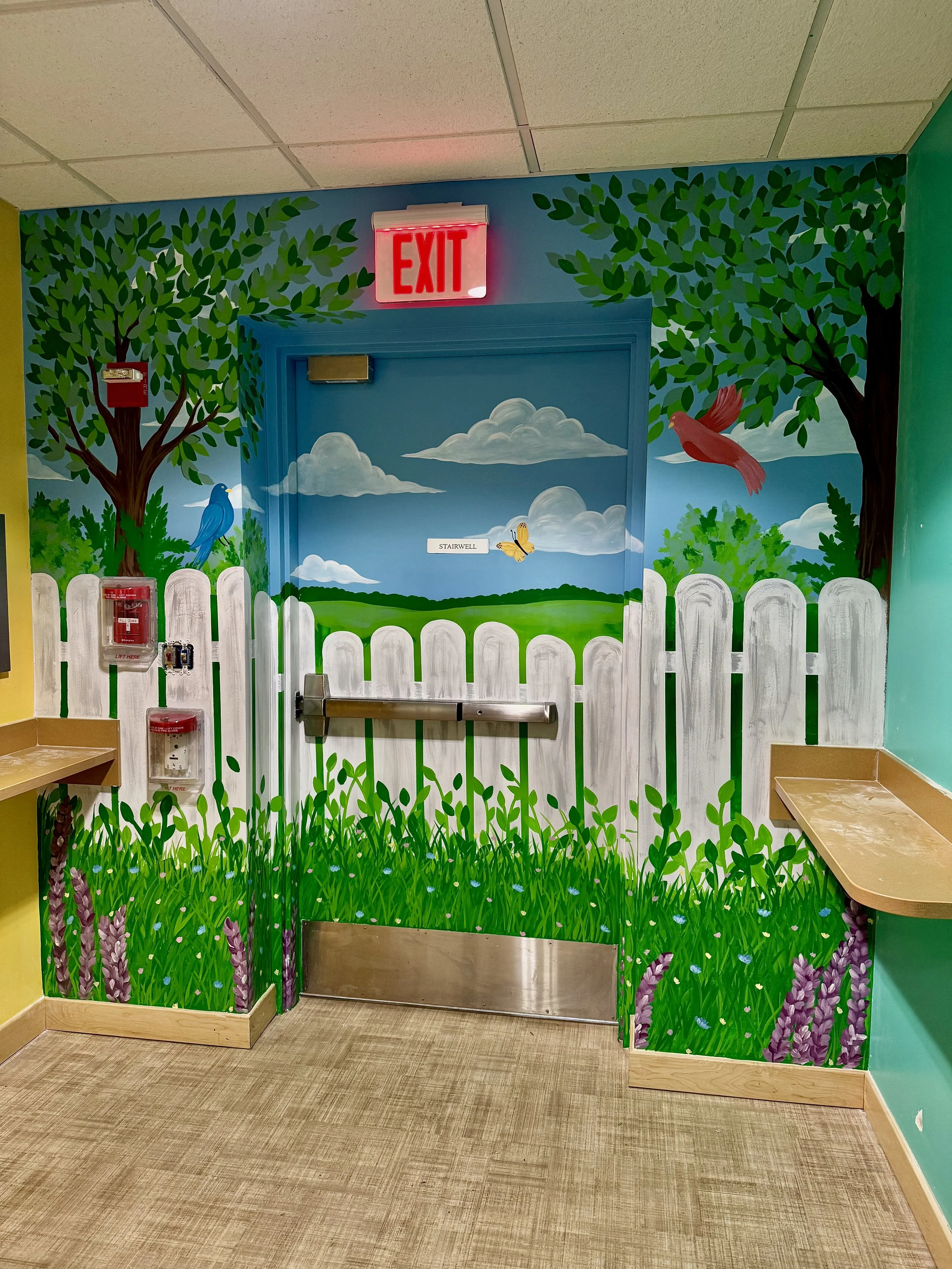 Colorful mural of a garden scene with trees, birds, clouds, grass, and flowers, surrounding a door with a 'STAIRWELL' sign, and a white picket fence painted on the wall.