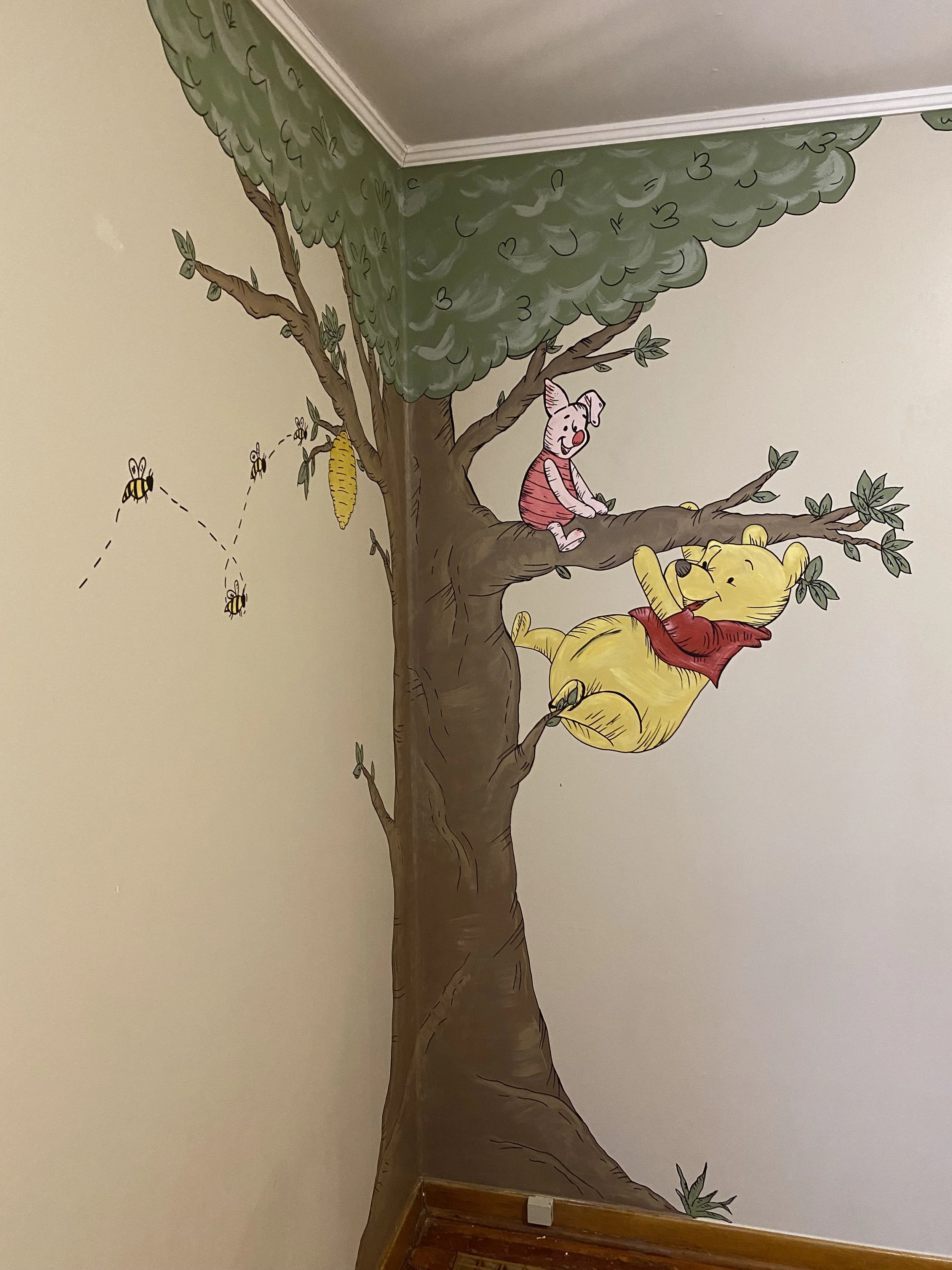 A nursery mural of Winnie the Pooh and Piglet sitting on a tree branch, surrounded by bees, leaves, and a honeycomb on a wall by Edmonton, AB artist Carly Marie Murals