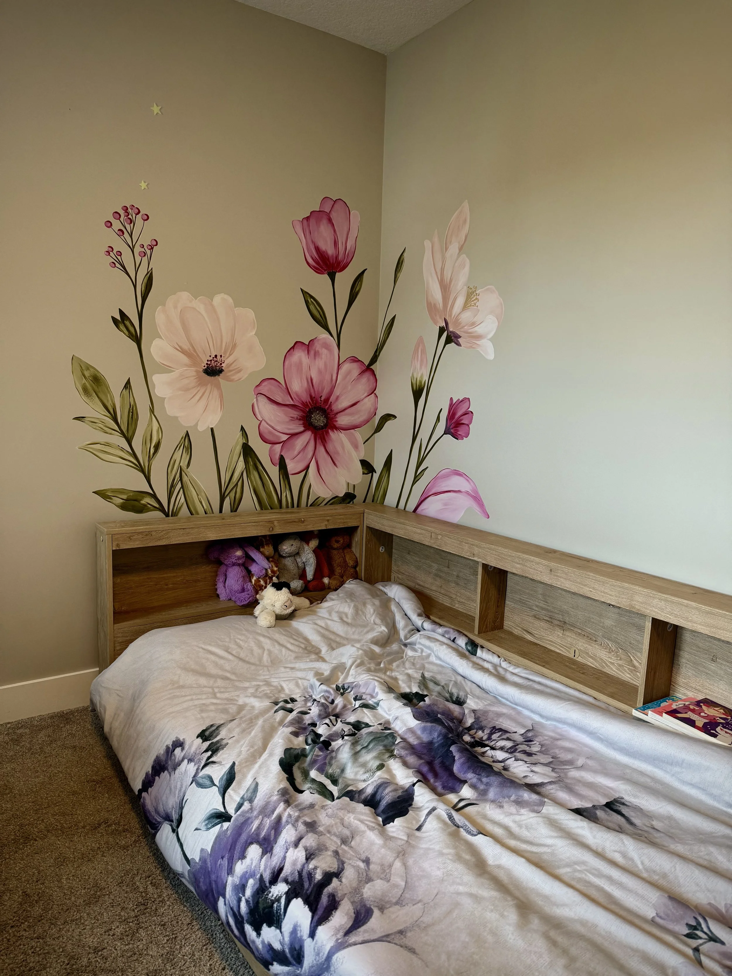 A children's bedroom with a wall mural of pink and white flowers with green leaves on the corner wall above children's bed. Mural painted by Edmonton, AB artist Carly Marie Murals 
