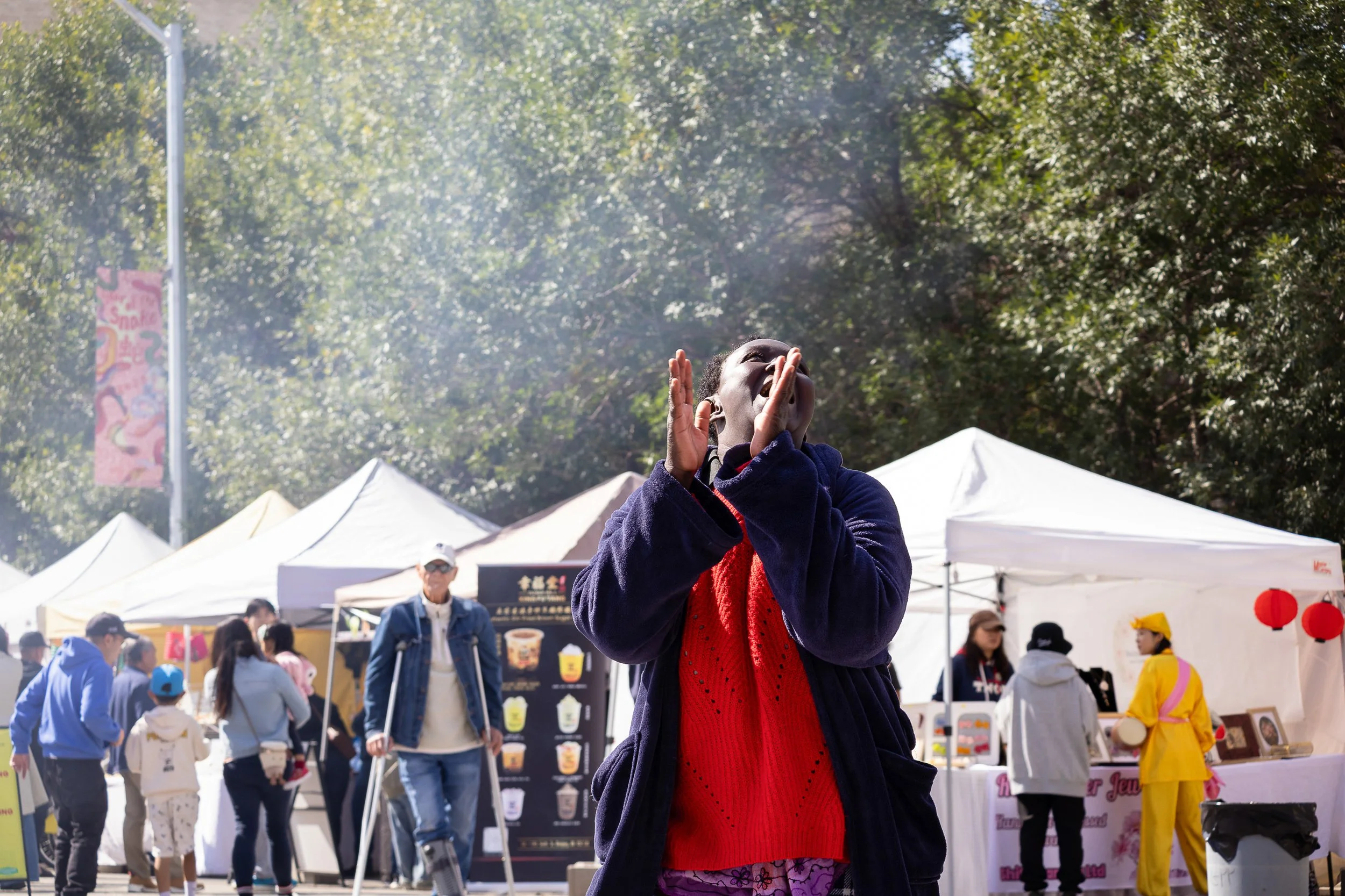 A woman calling out with hands cupped at an outdoor festival with tents and crowd in the background.