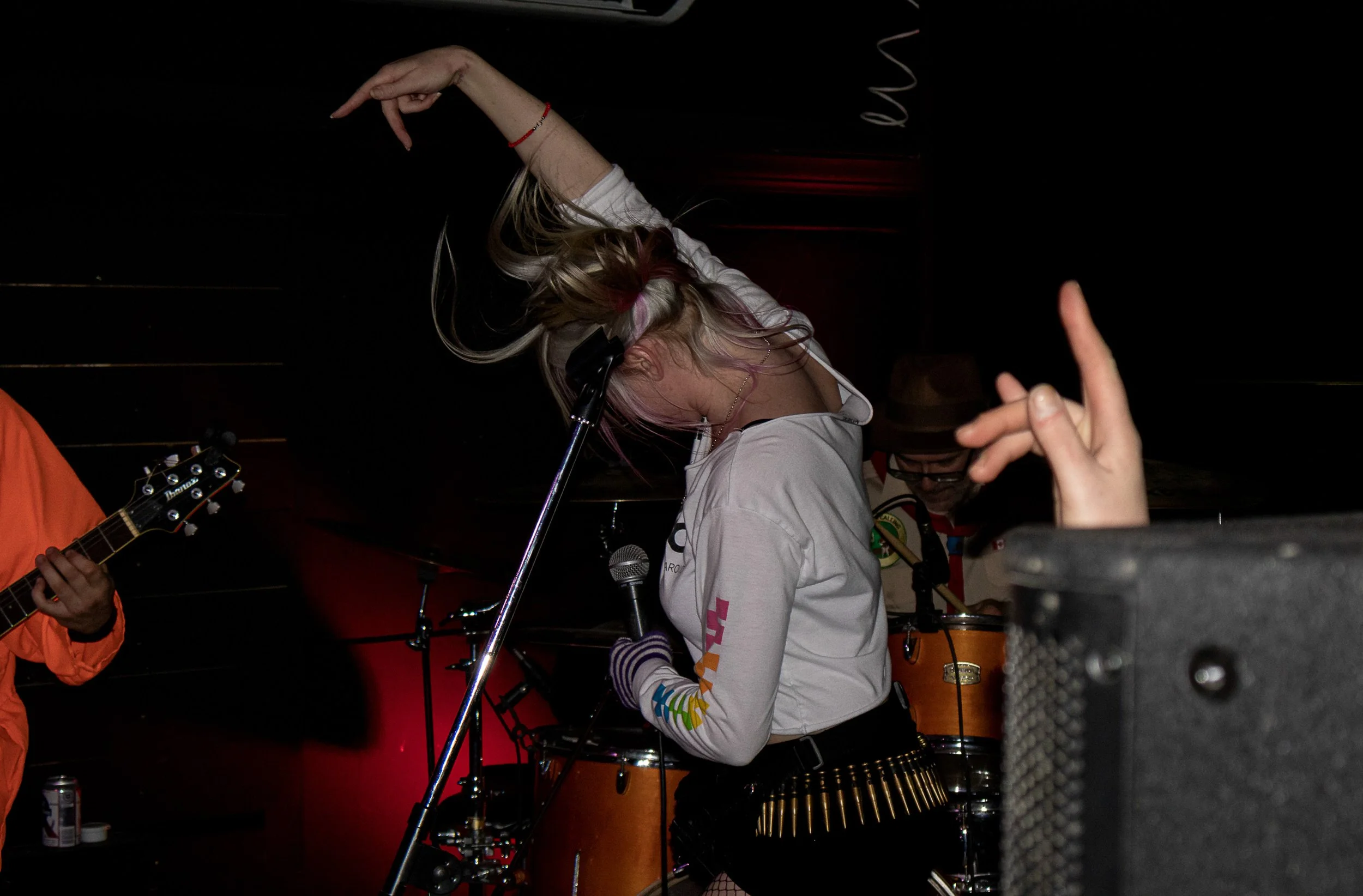 A punk rock singer with raised arm performs energetically at a small live music show in Winnipeg, band members and stage light behind her.