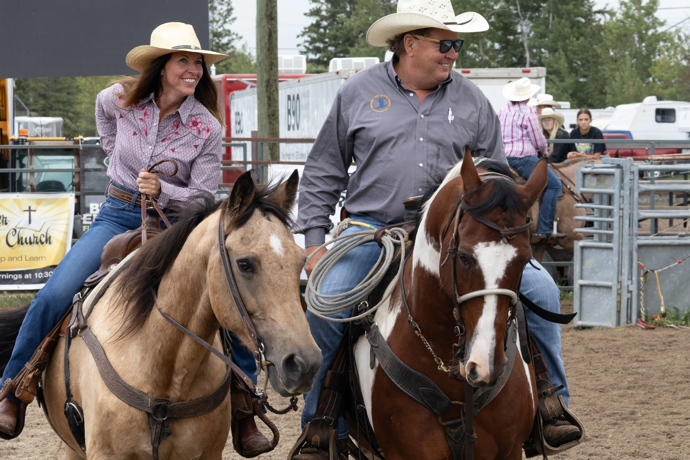 Two riders in cowboy hats sit on horseback at a rodeo arena, smiling and holding reins, with livestock pens and spectators behind them.