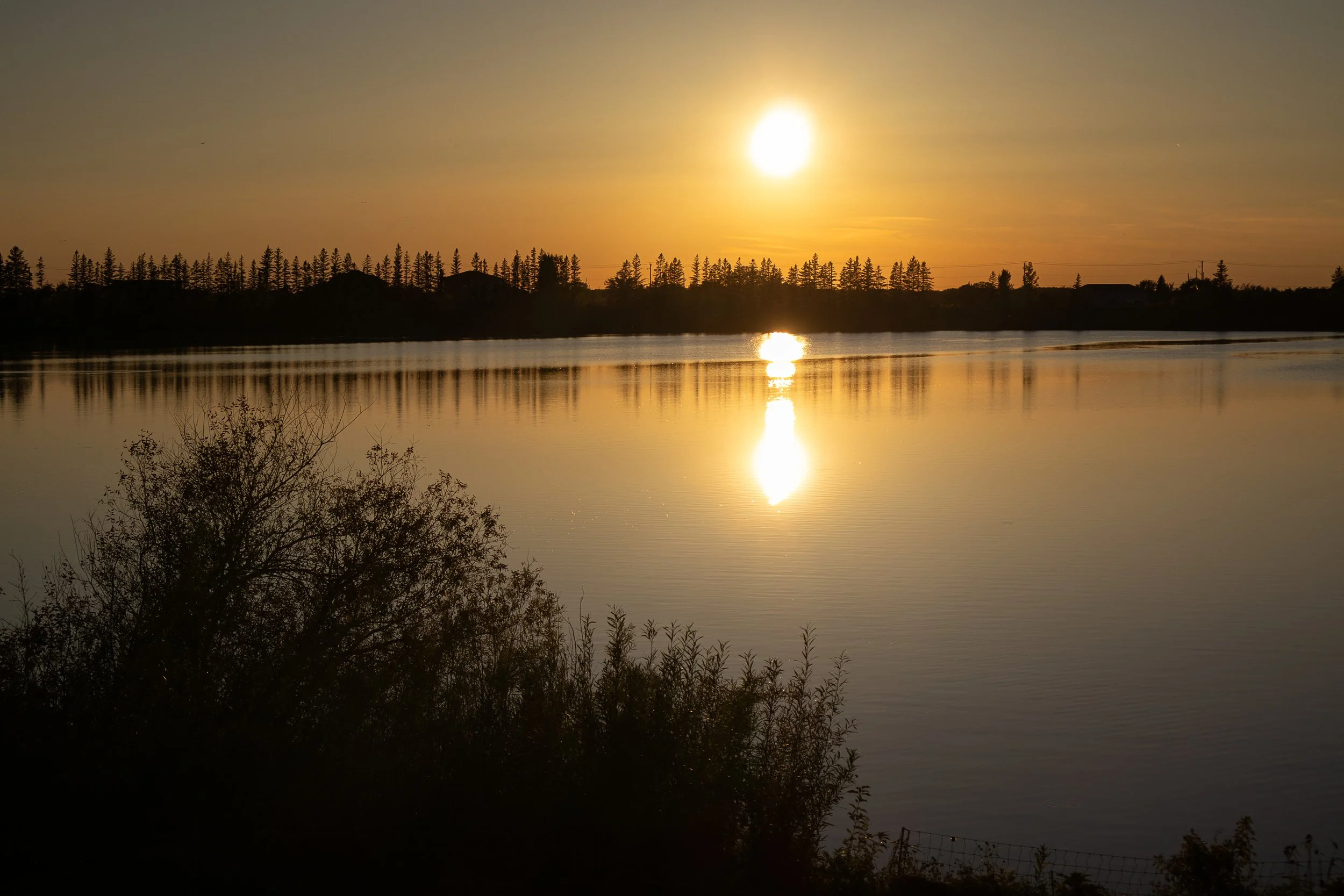 A bright orange sun hangs above a calm lake at dusk, its reflection stretching across the water toward a dark tree line on the horizon.