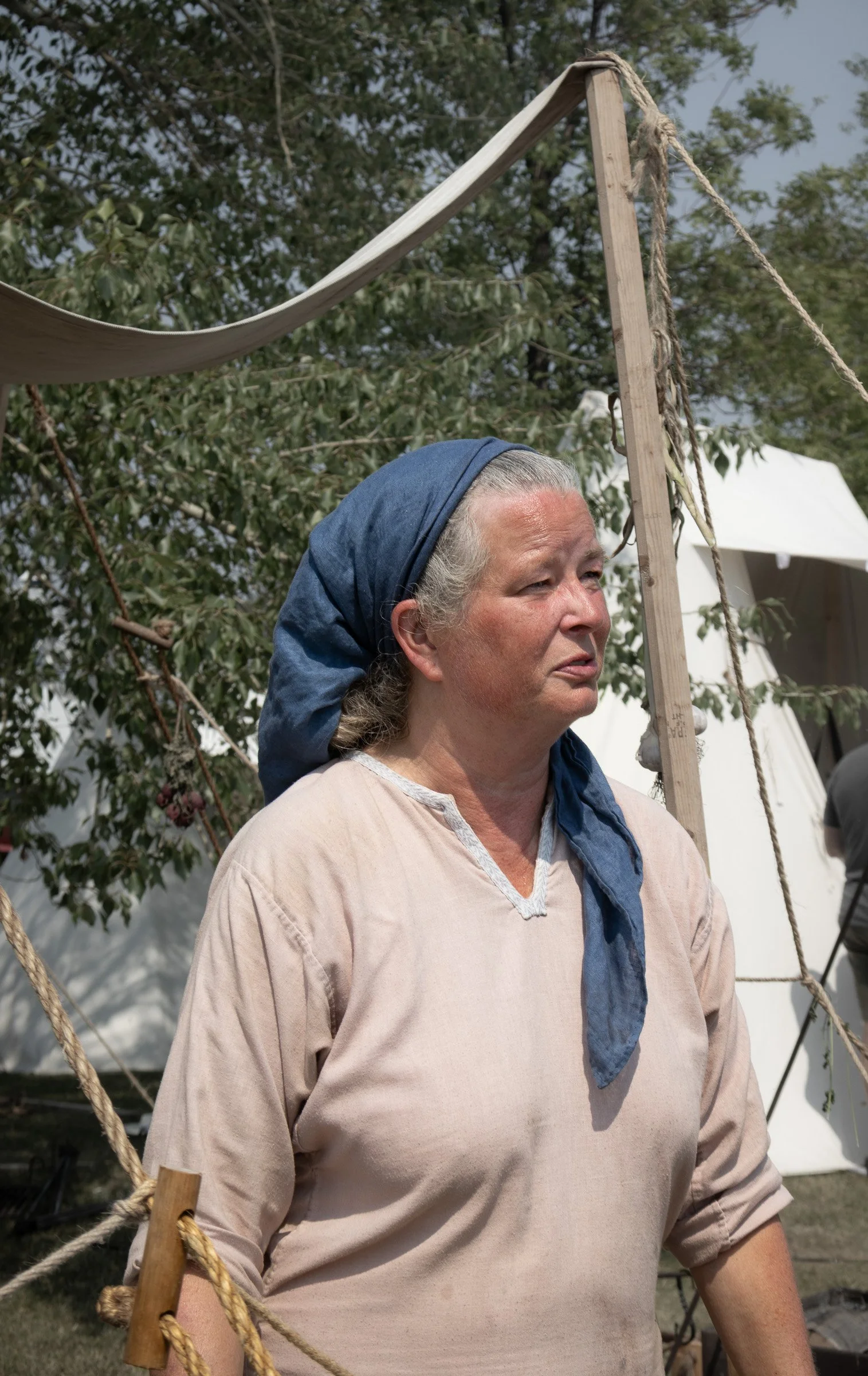 A woman wearing a blue headscarf and plain long-sleeved shirt stands outdoors near canvas tents and wooden poles, looking off to the ride, village of Icelanders in Gimli.