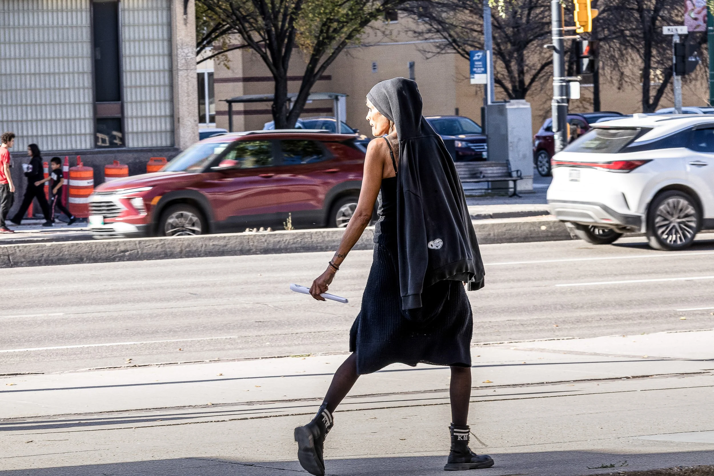 A woman dressed in black crosses a prairie city street, sunlight catching her as traffic moves in the background.