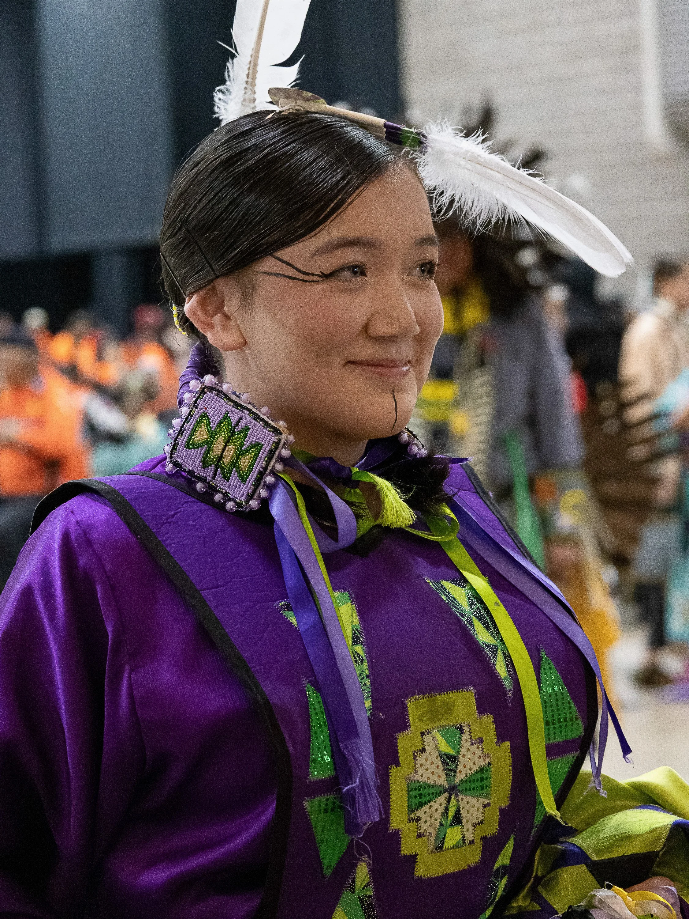 Indigenous participants in traditional regalia-a woman in a purple beaded dress with feather adornment and a man wearing a feathered headdress and beaded accessories-at a cultural gathering.