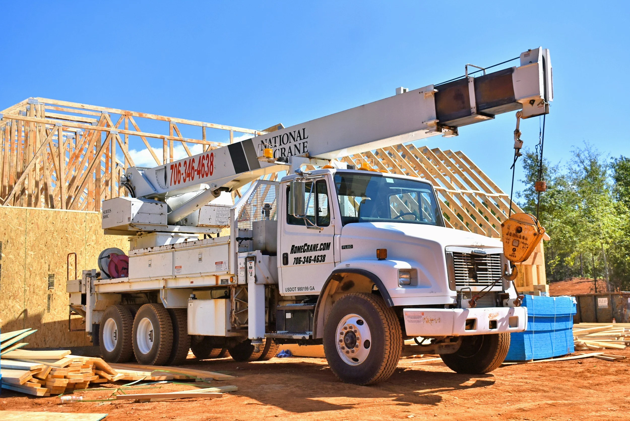 A white crane truck with a mounted extendable boom and hook at a construction site. Wooden framing of a building under construction is visible in the background, along with stacks of lumber and construction materials on the ground.