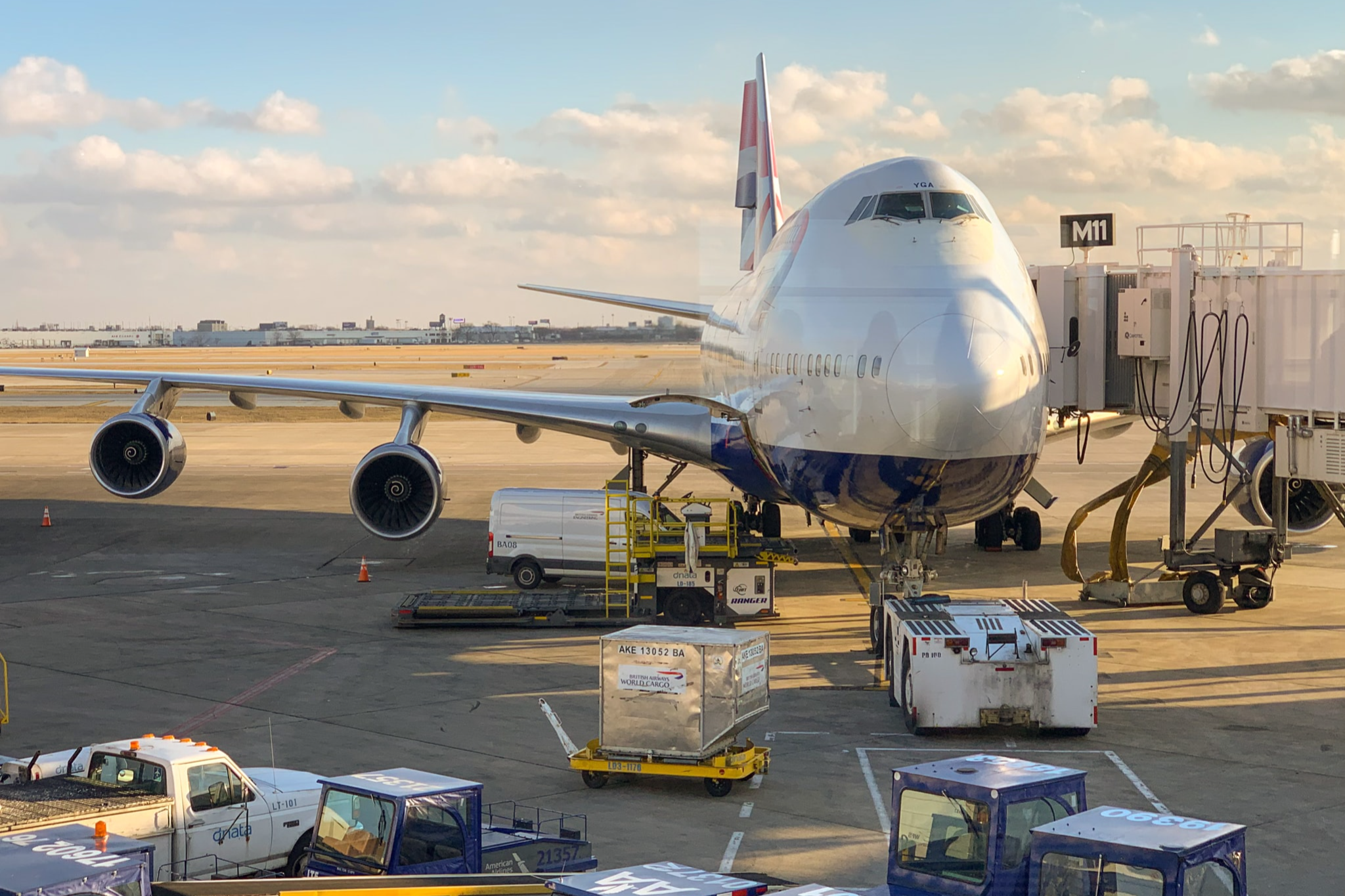 Boeing-style commercial airplane parked at gate with jet bridge, luggage carts, and service vehicles on tarmac at airport.